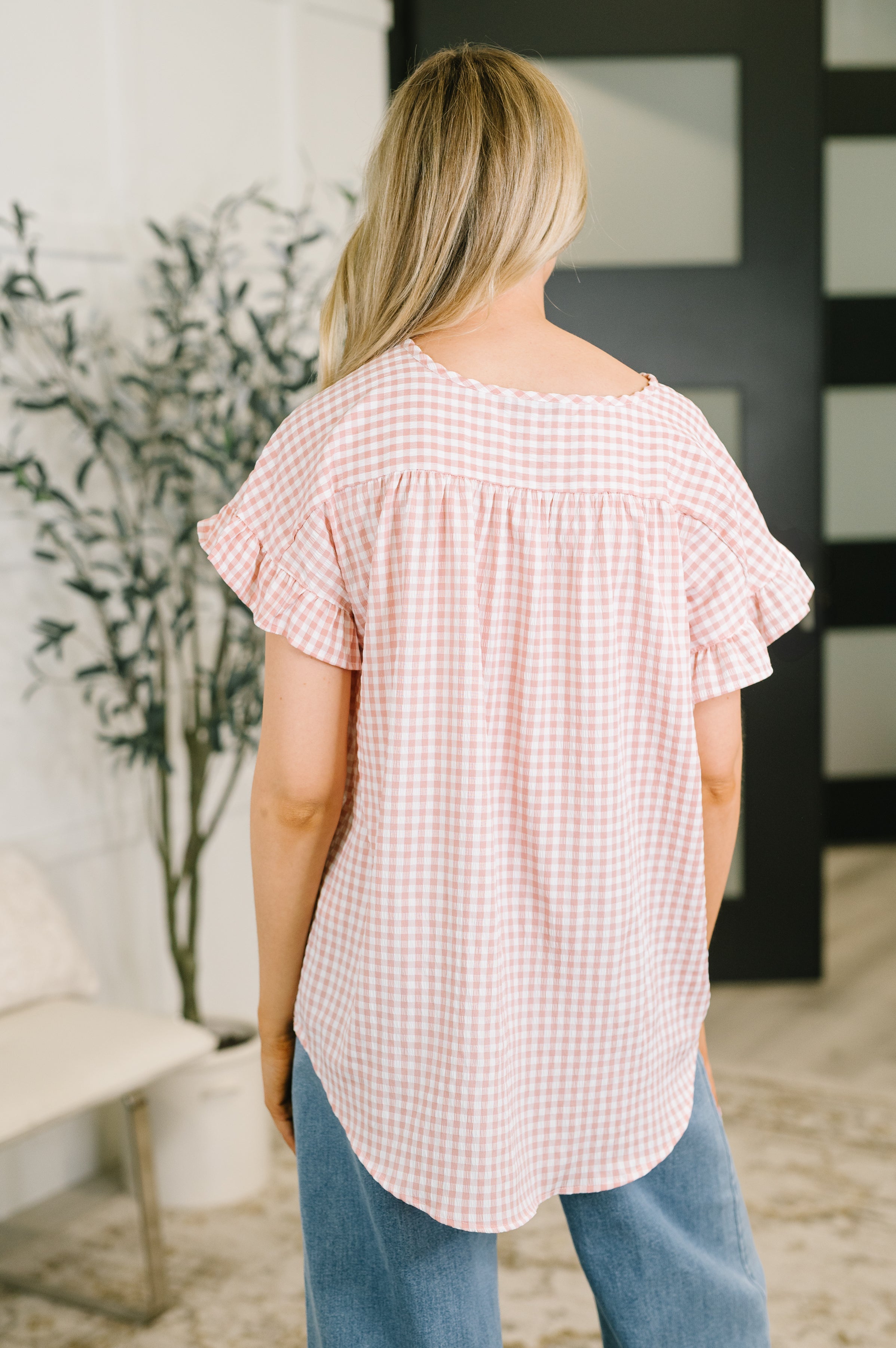 Back view of woman wearing a dusty rose gingham blouse with ruffled sleeves and curved hem indoors