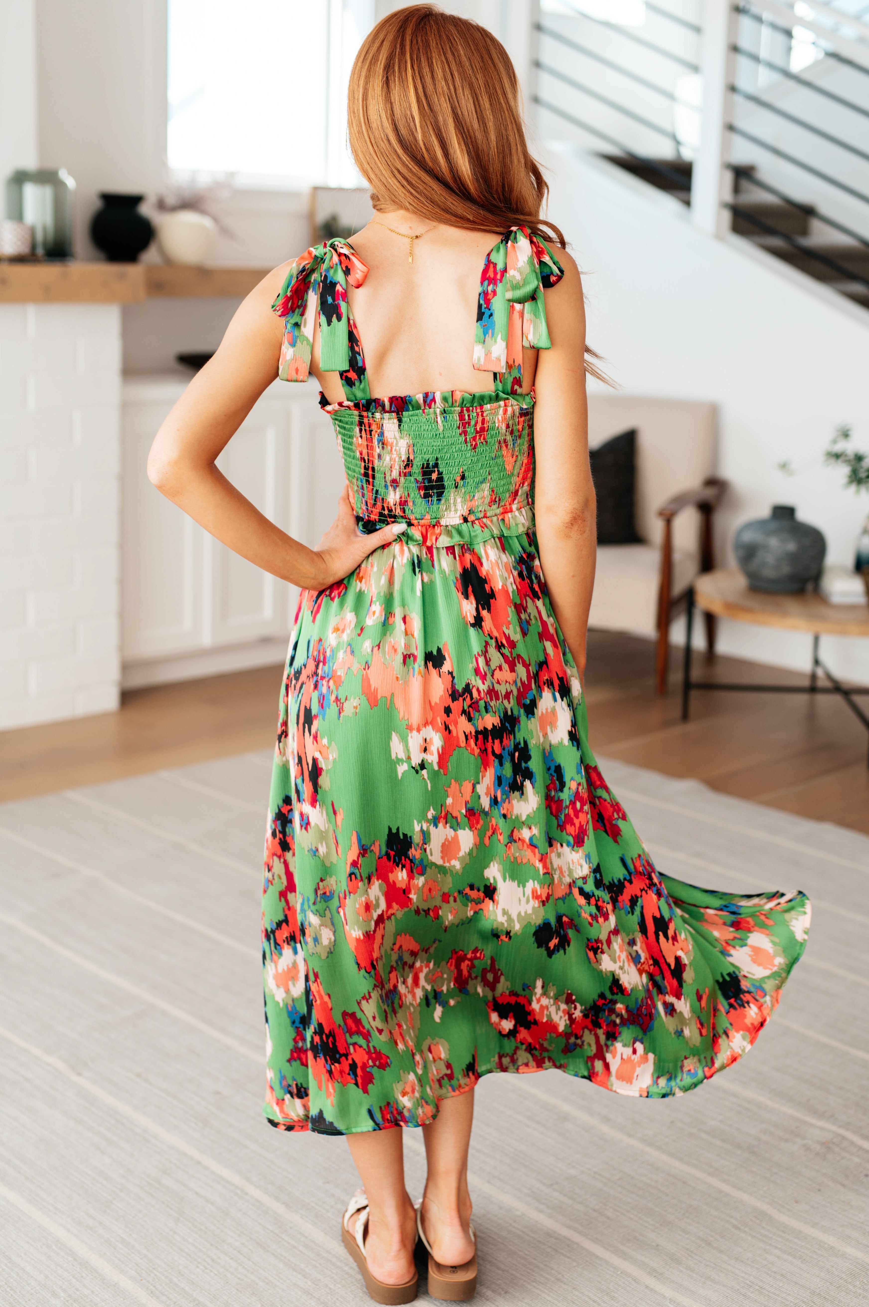 Woman indoors wearing green floral print dress with smocked back and tie straps, view from behind showing flowing skirt