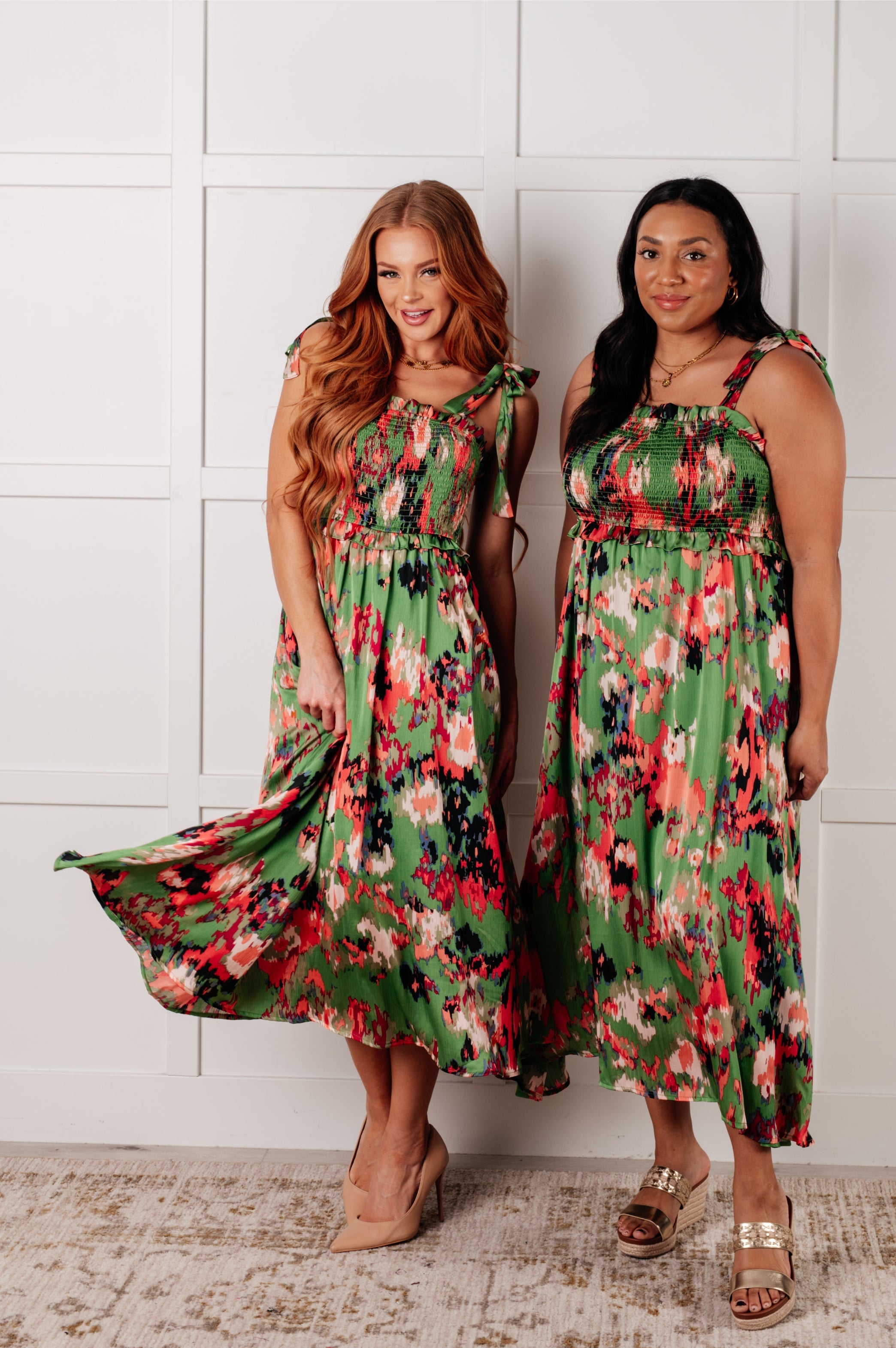Two women wearing green floral smocked dresses with shoulder ties posing against white paneled wall