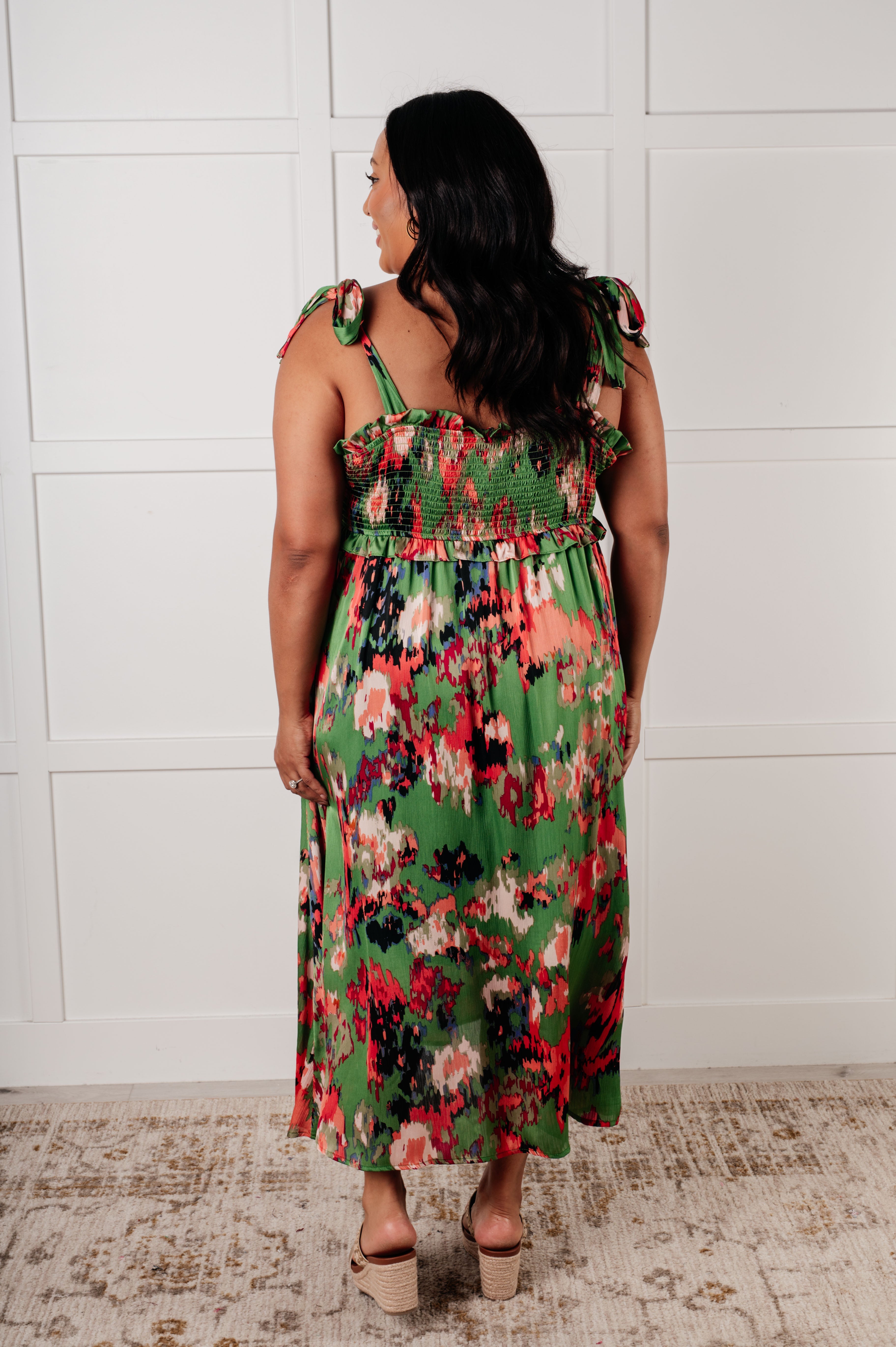 Woman wearing green floral midi dress with tie straps and smocked back standing on patterned rug facing away