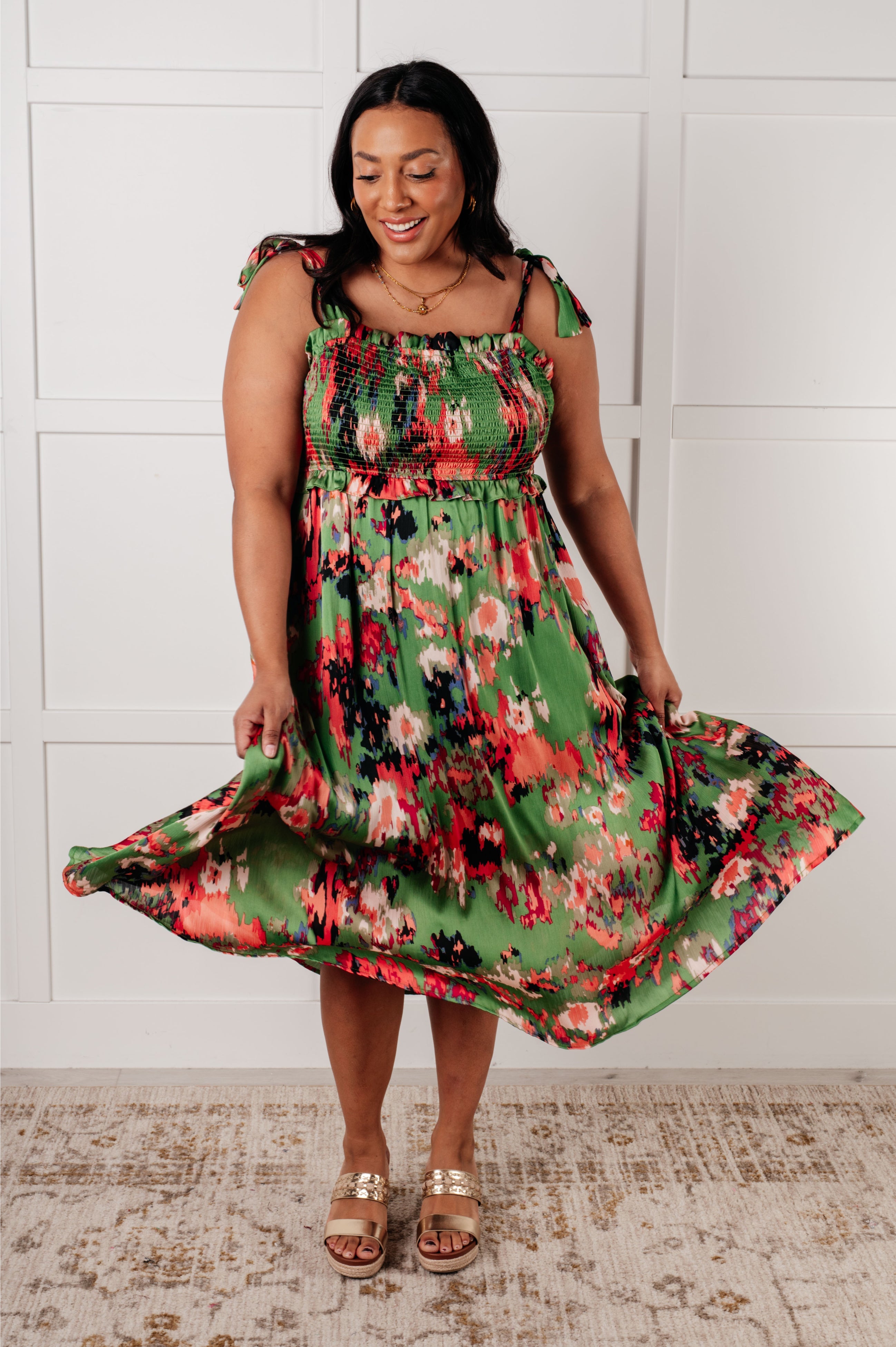 Woman twirling in green floral smocked dress with shoulder ties and flowing skirt, standing indoors front view