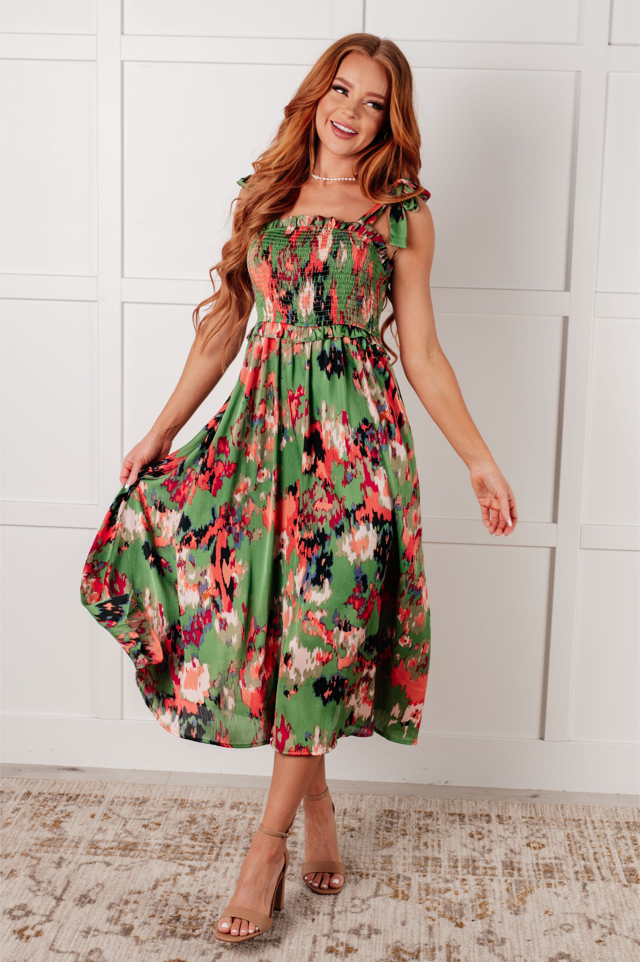 Woman with long wavy hair wearing green floral A Little While Longer Dress with smocked bodice and tie shoulder straps standing indoors