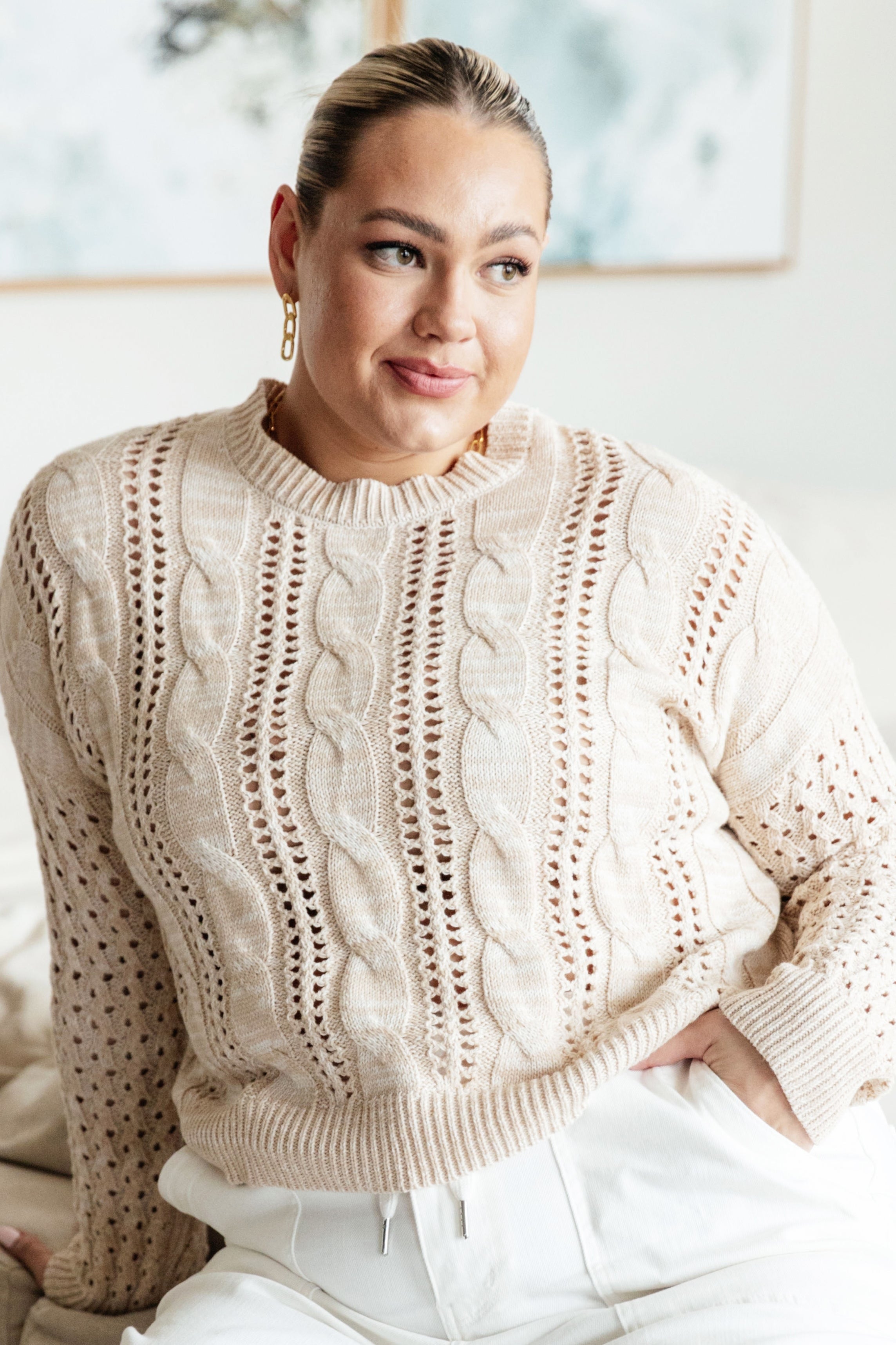 Woman with slicked-back hair wearing cream cable knit sweater with open stitch details sitting indoors front view