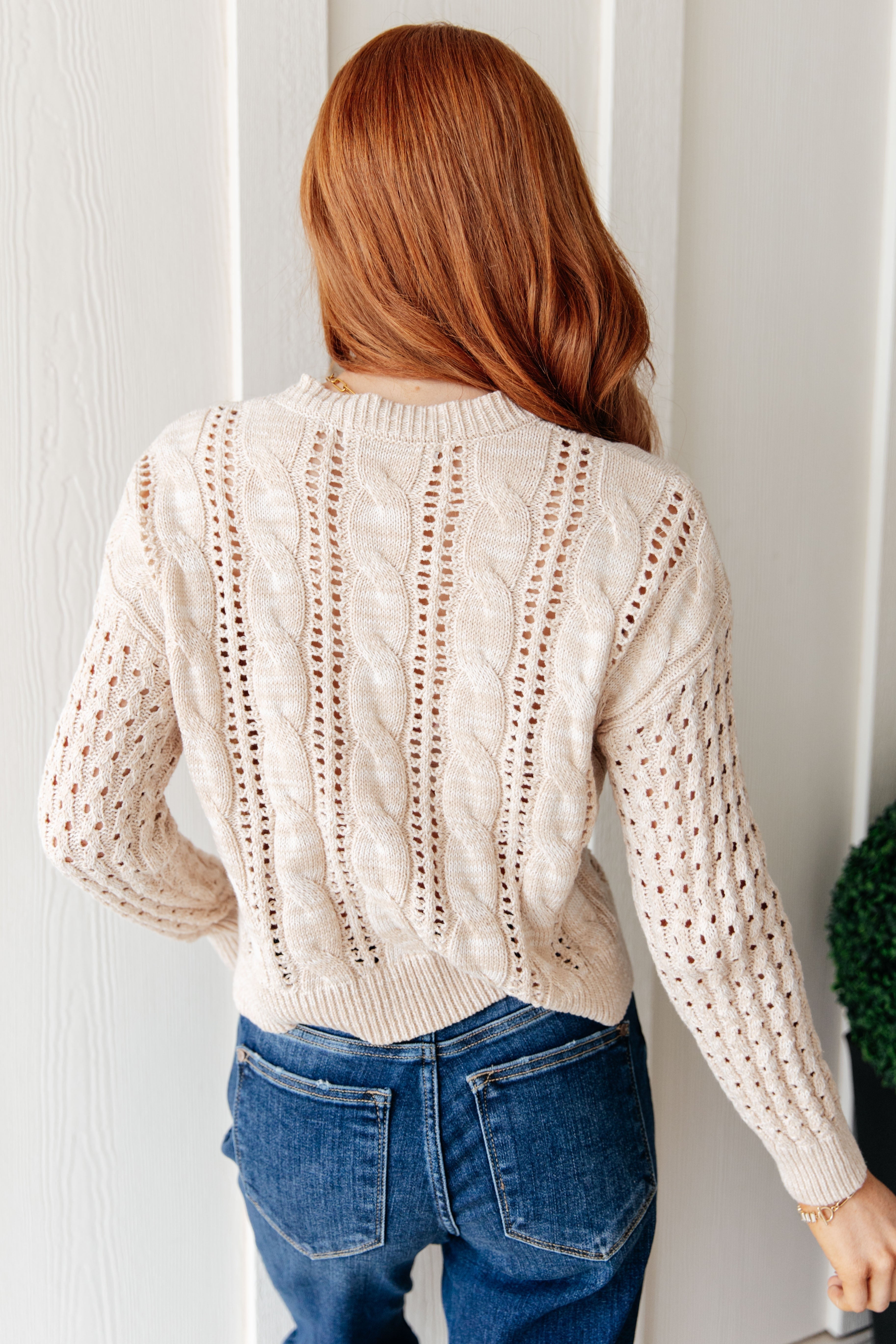 Red-haired woman seen from behind wearing cream cable knit sweater with openwork details and blue jeans indoors