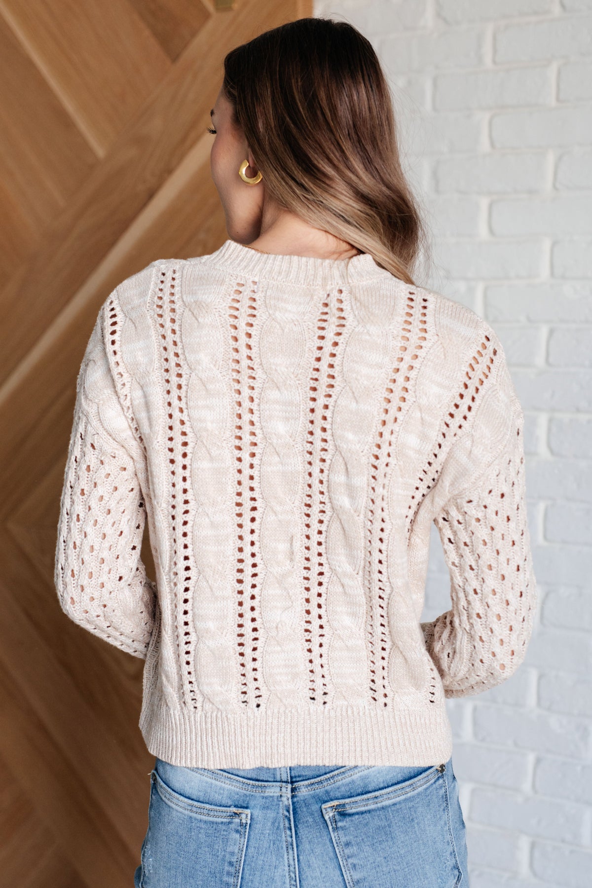 Back view of a woman wearing a cream cable knit sweater with openwork details and blue jeans indoors