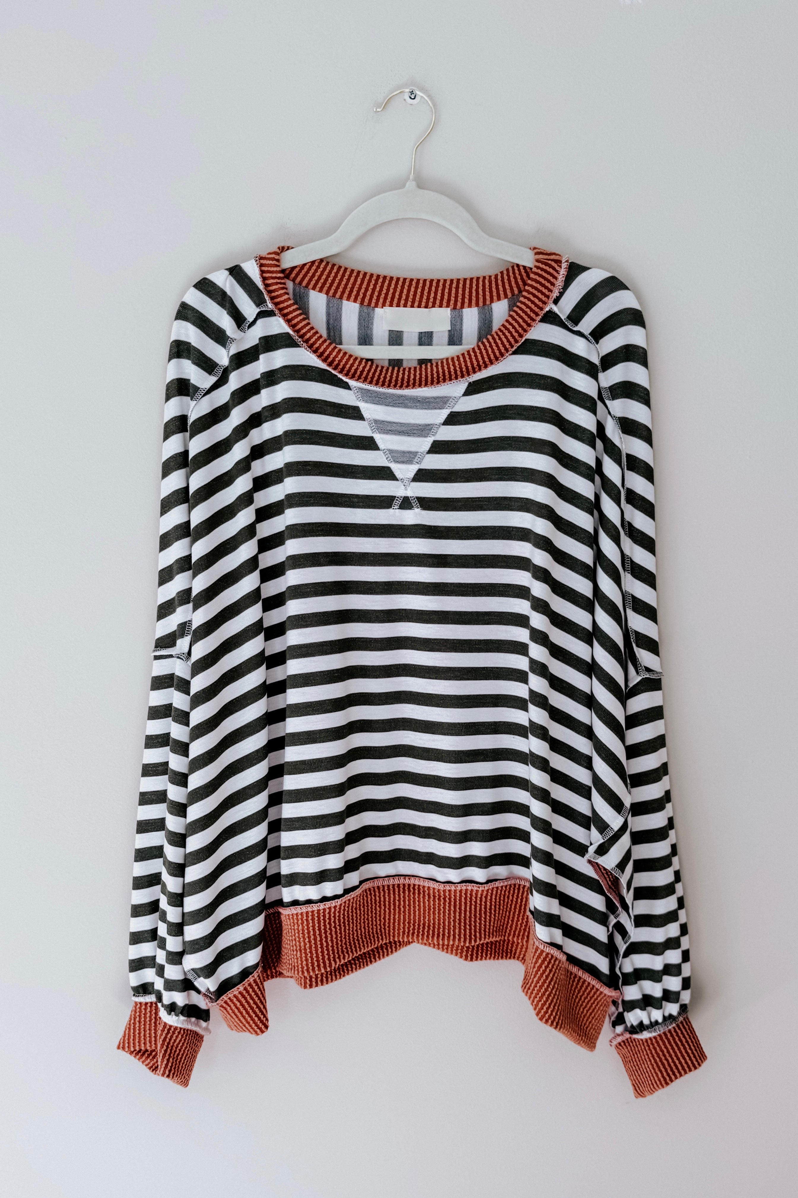 Black and white striped crew neck pullover with rust-colored ribbed collar, cuffs, and hem hanging on white hanger against gray wall