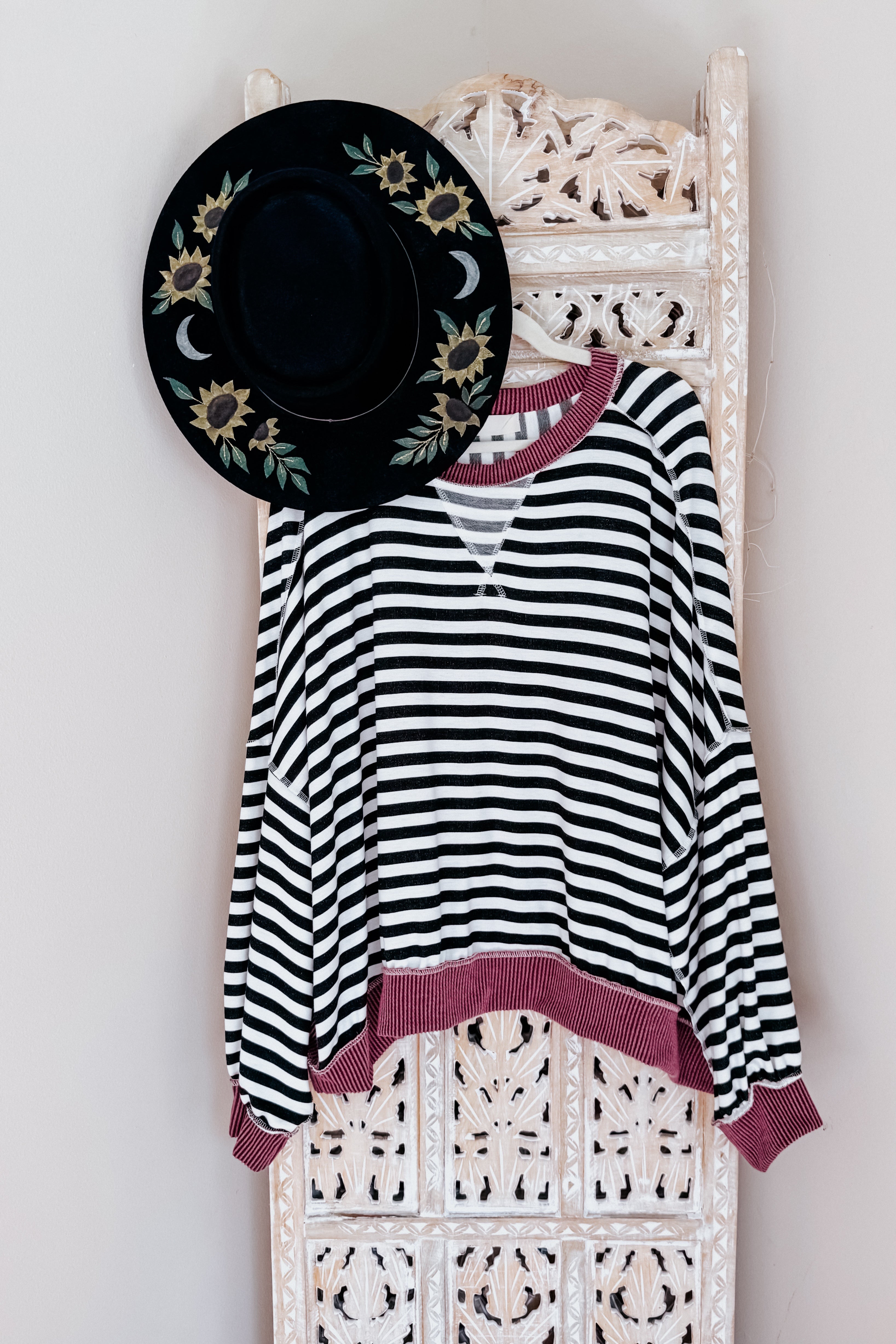 Black and white striped crew neck pullover with wine ribbed trim hanging on carved white wooden screen with black floral hat