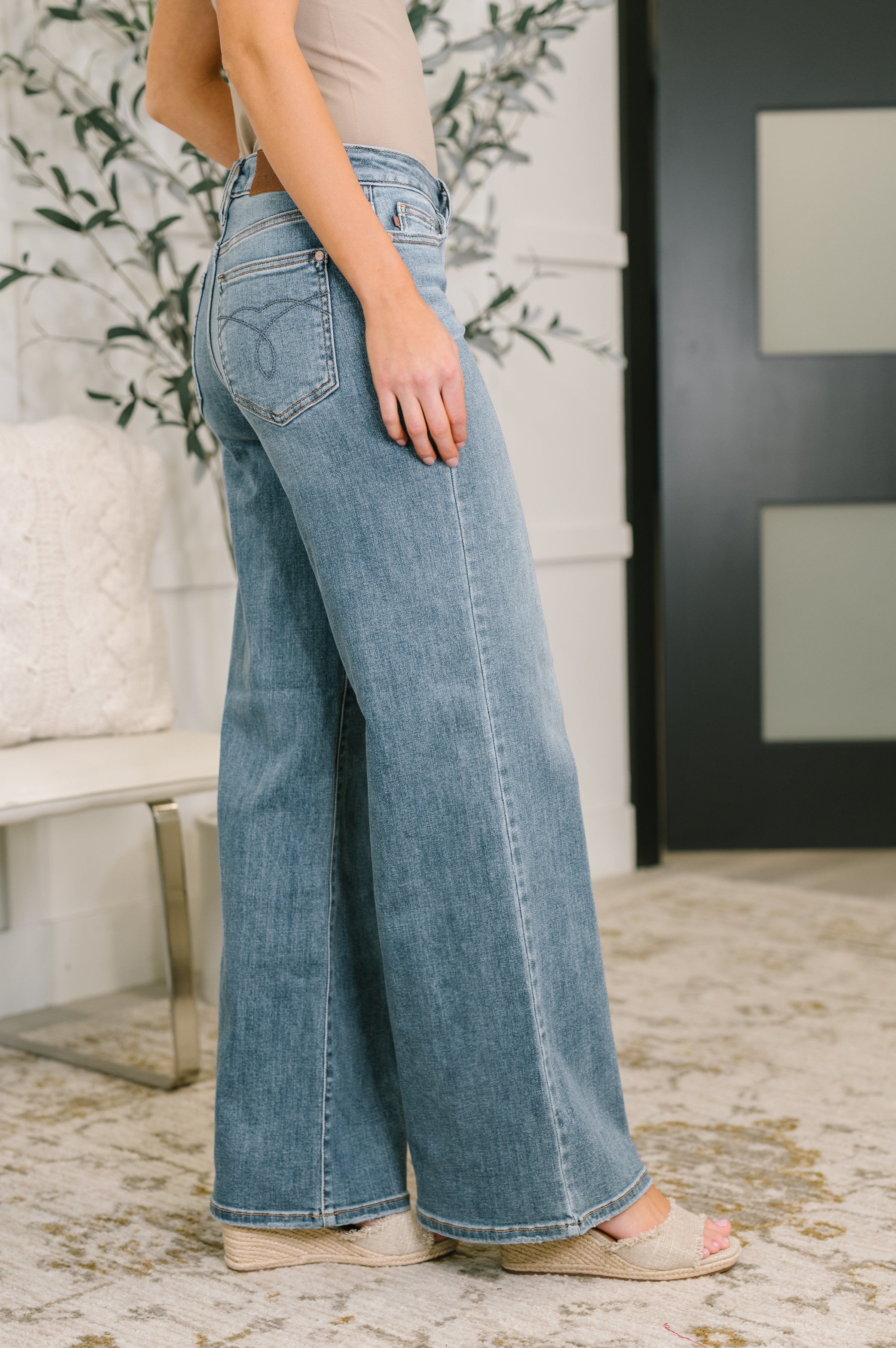 Side view of Adrian mid rise wide leg jeans in medium wash denim worn with neutral top and wedge sandals indoors