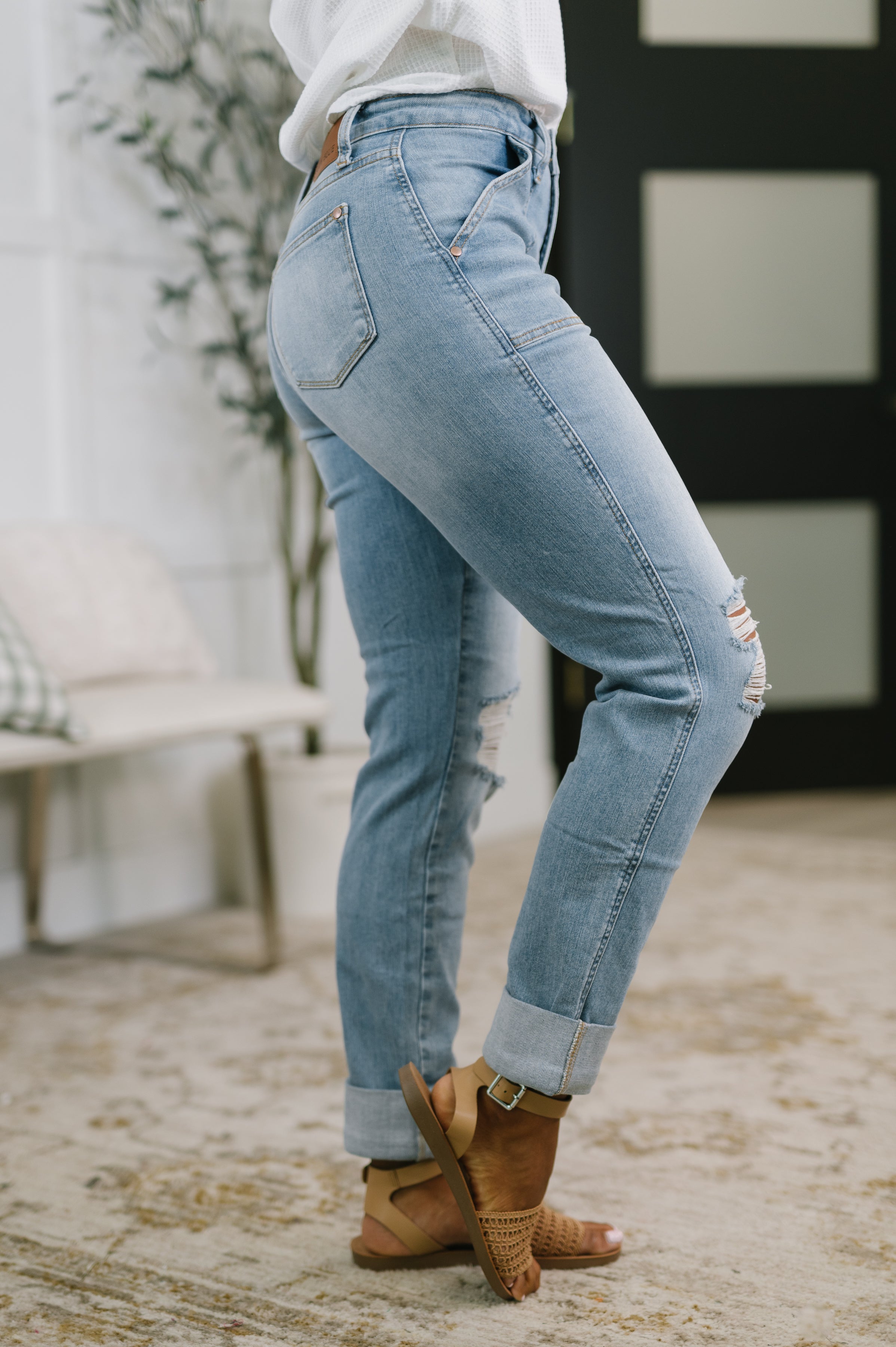 Side view of woman wearing light blue distressed boyfriend jeans with rolled cuffs and tan sandals indoors