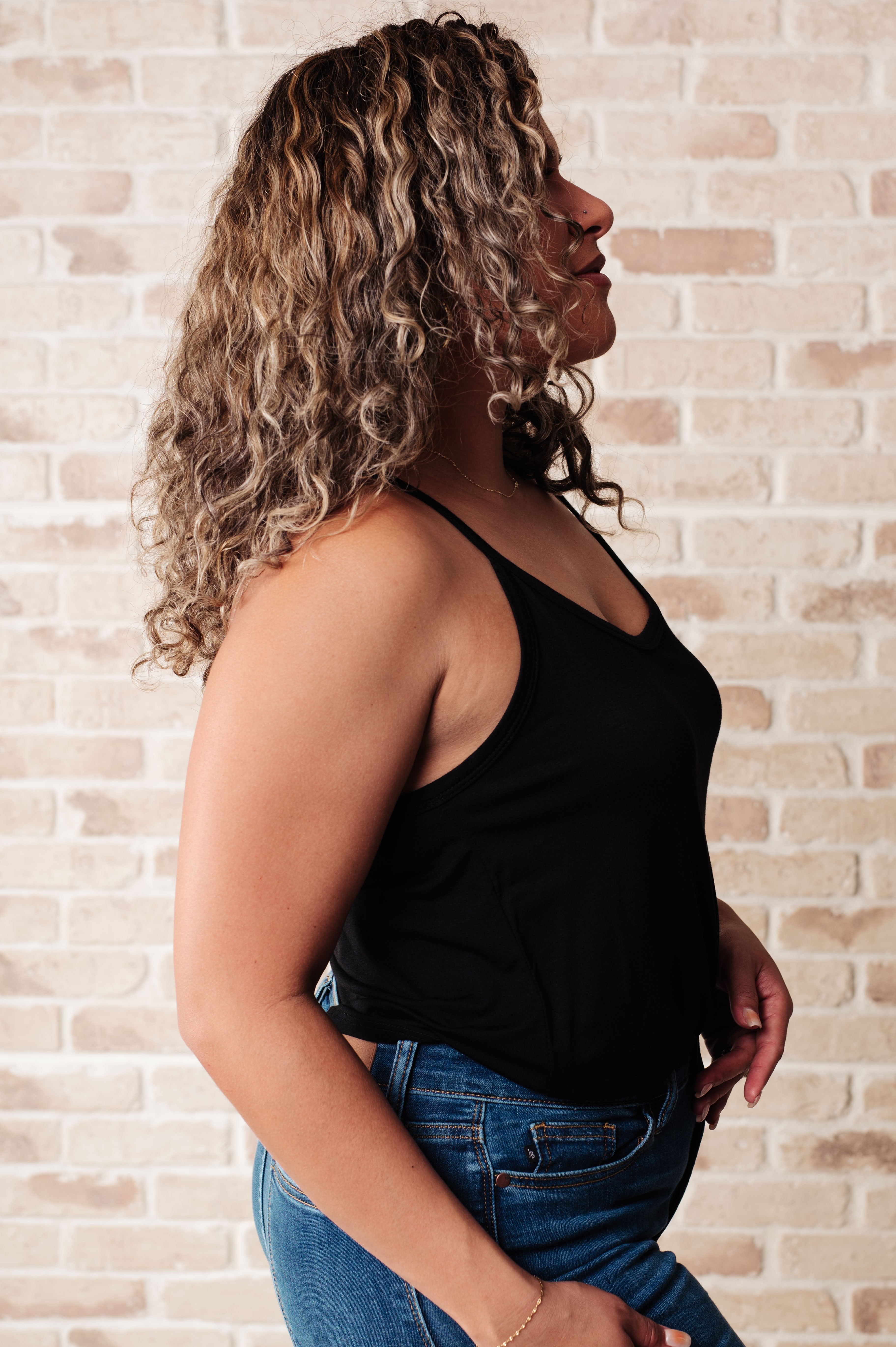 Side profile of woman wearing black Ain't No Mountain High Enough tank top paired with blue jeans against brick wall
