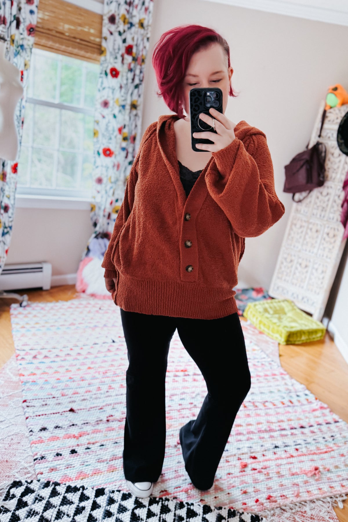 Woman taking a mirror selfie in a rust textured hooded henley sweater with half button closure black pants and white shoes indoors