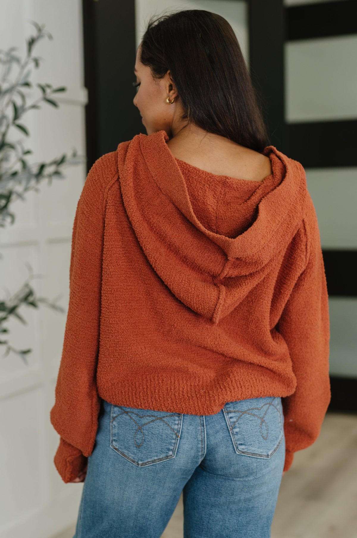 Woman viewed from behind wearing a rust textured hooded sweater with relaxed sleeves and blue jeans indoors