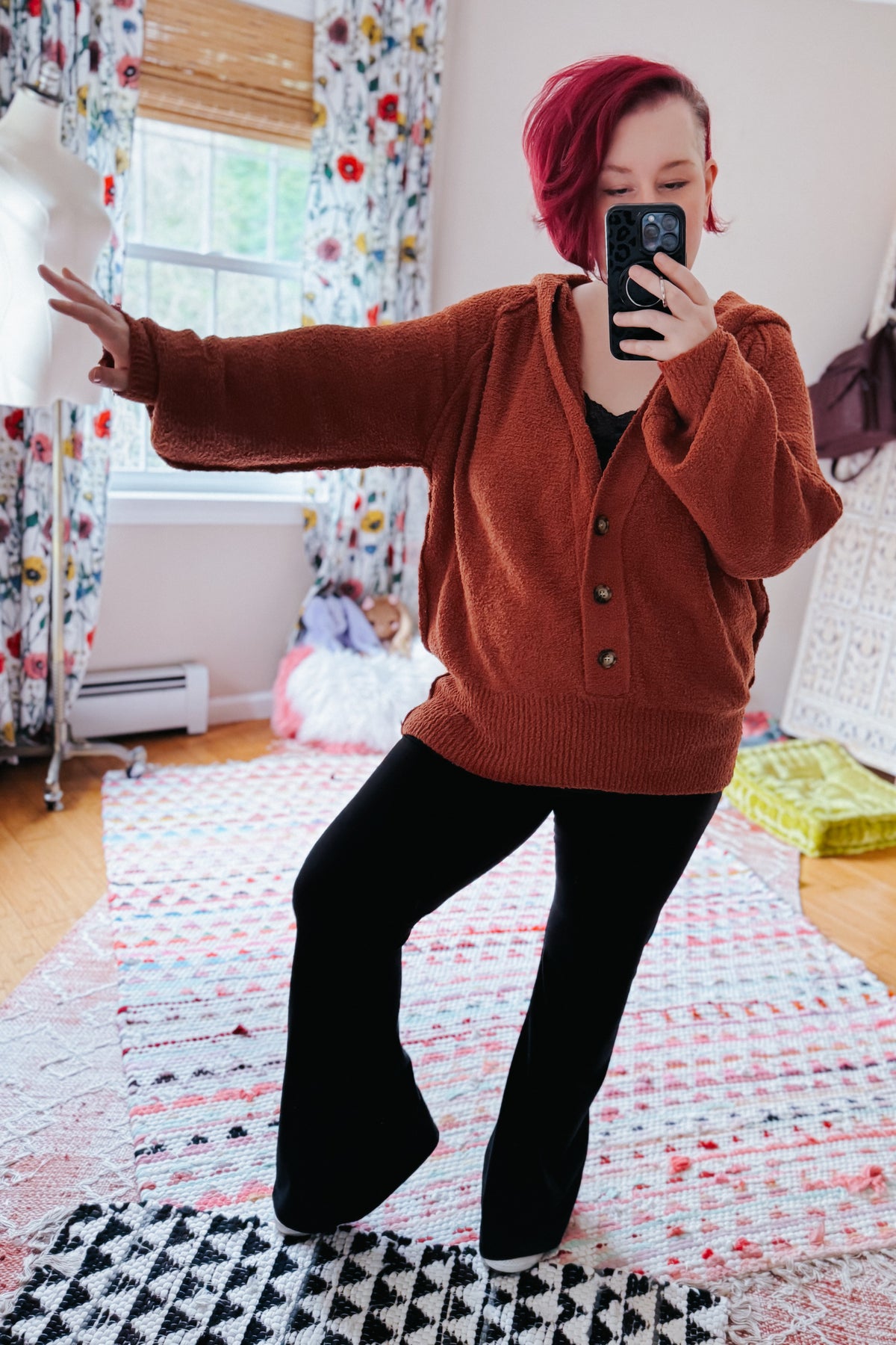 Woman taking a mirror selfie in a rust textured hooded henley sweater with half button closures and black pants indoors