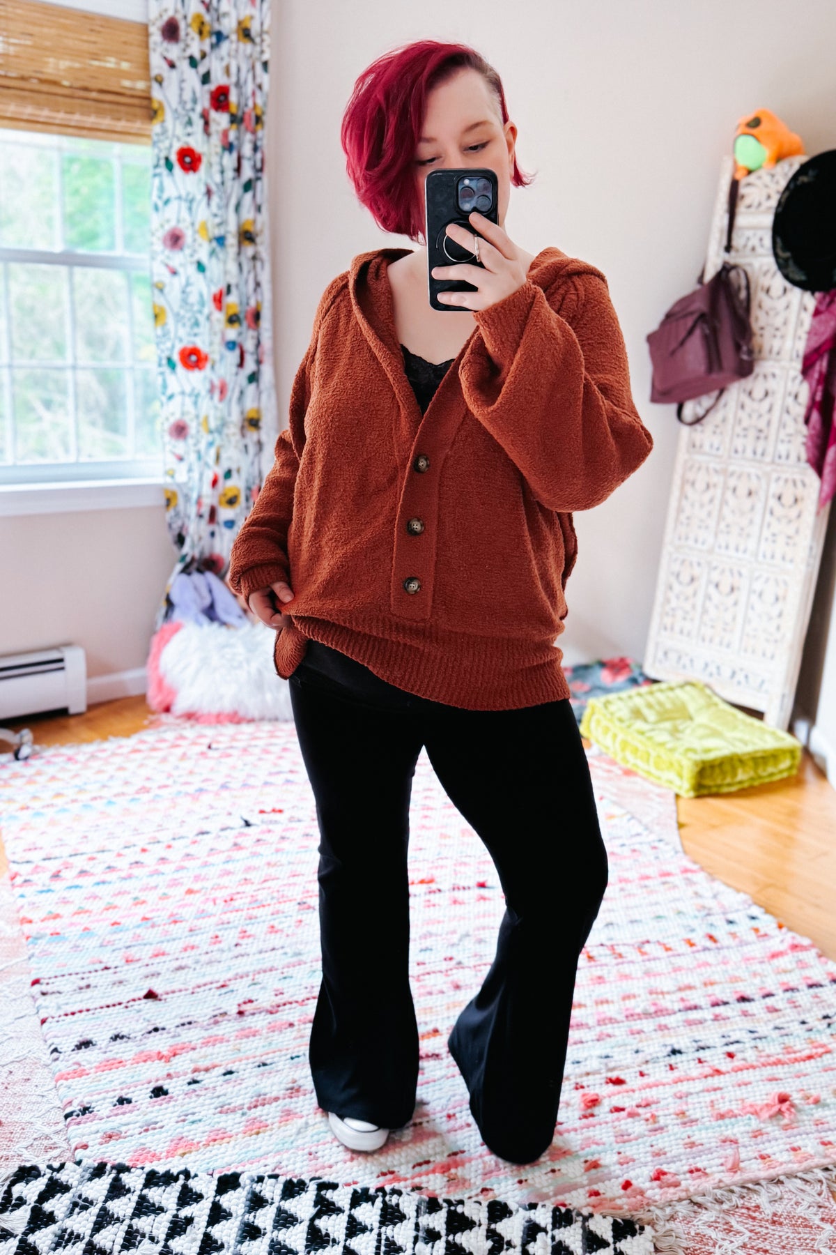 Woman taking a mirror selfie in a rust textured hooded henley sweater with button details and black pants indoors