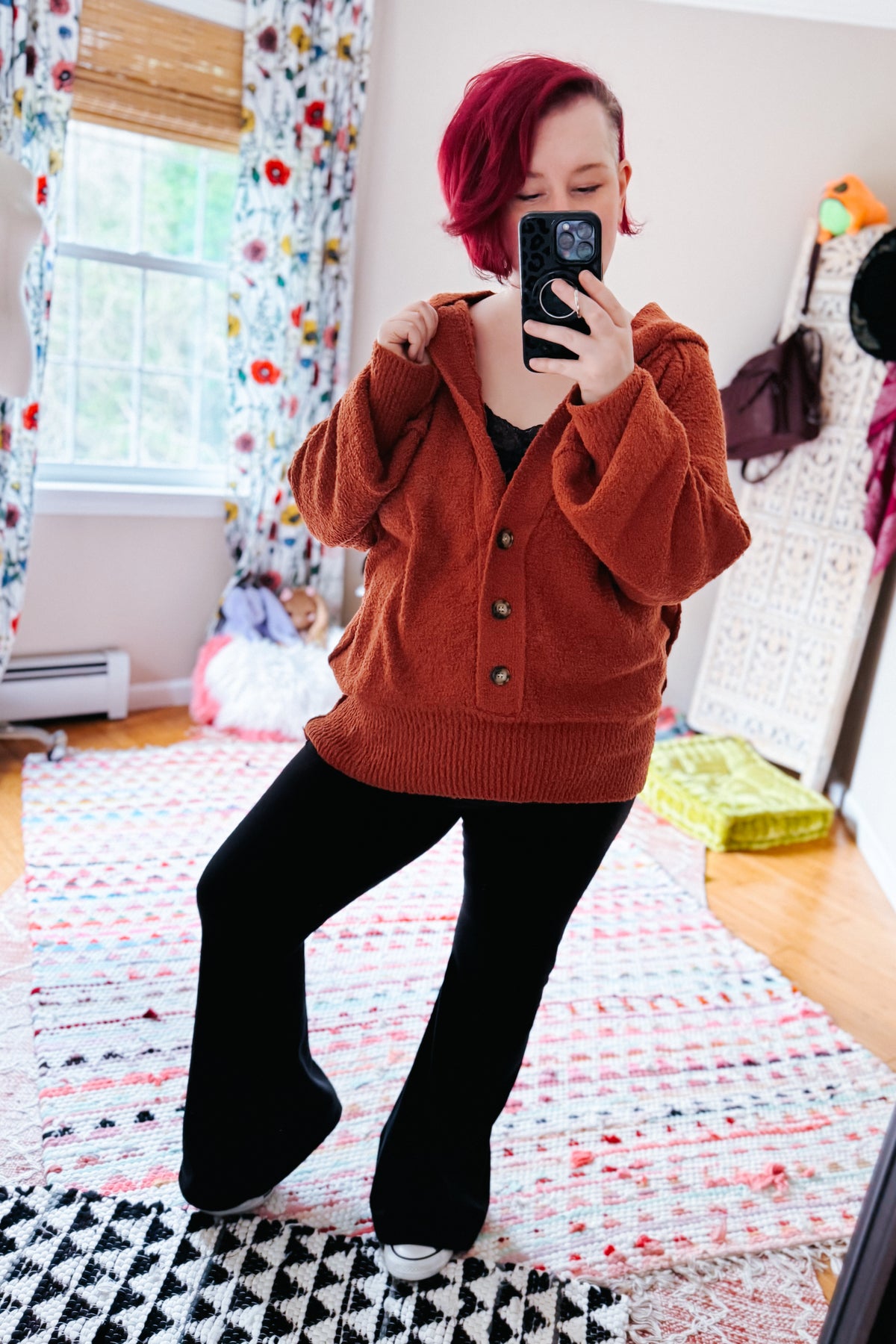 Woman posing for a mirror selfie in a rust textured hooded henley sweater with button details, black pants, and white sneakers indoors