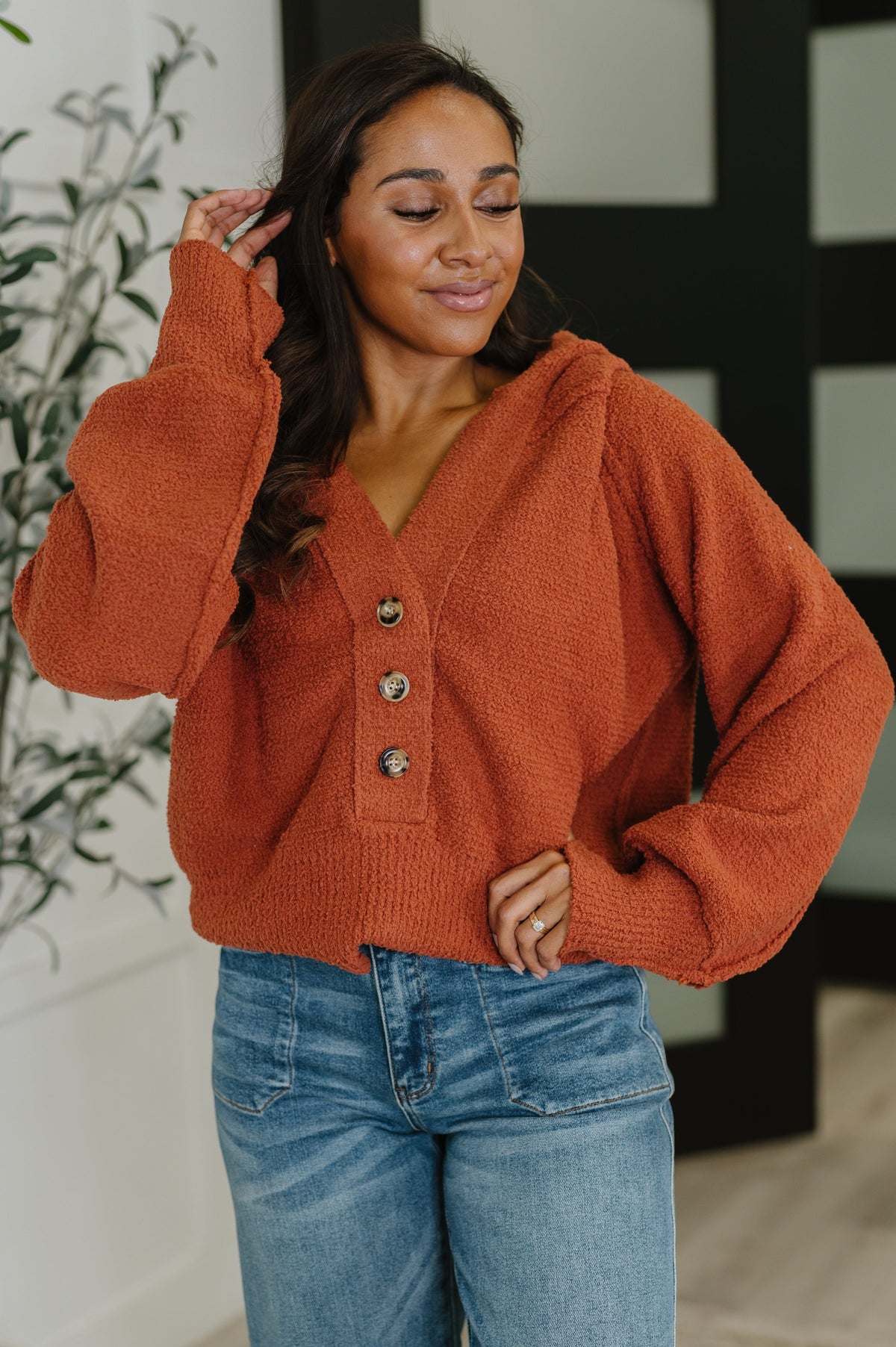 Woman modeling a rust textured hooded henley sweater with V neckline and half button front paired with blue jeans indoors