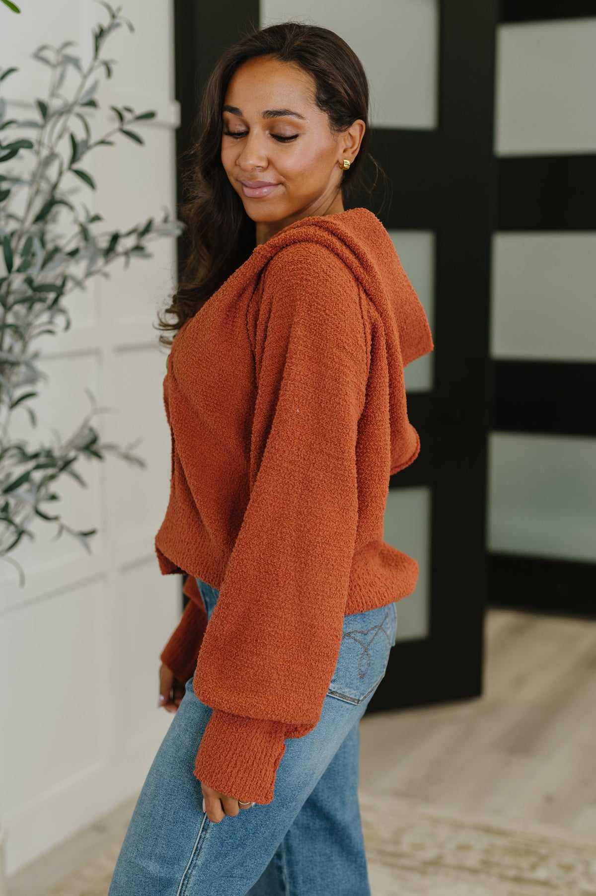 Woman modeling a rust textured hooded henley sweater with exposed seams and blue jeans in a bright modern home interior