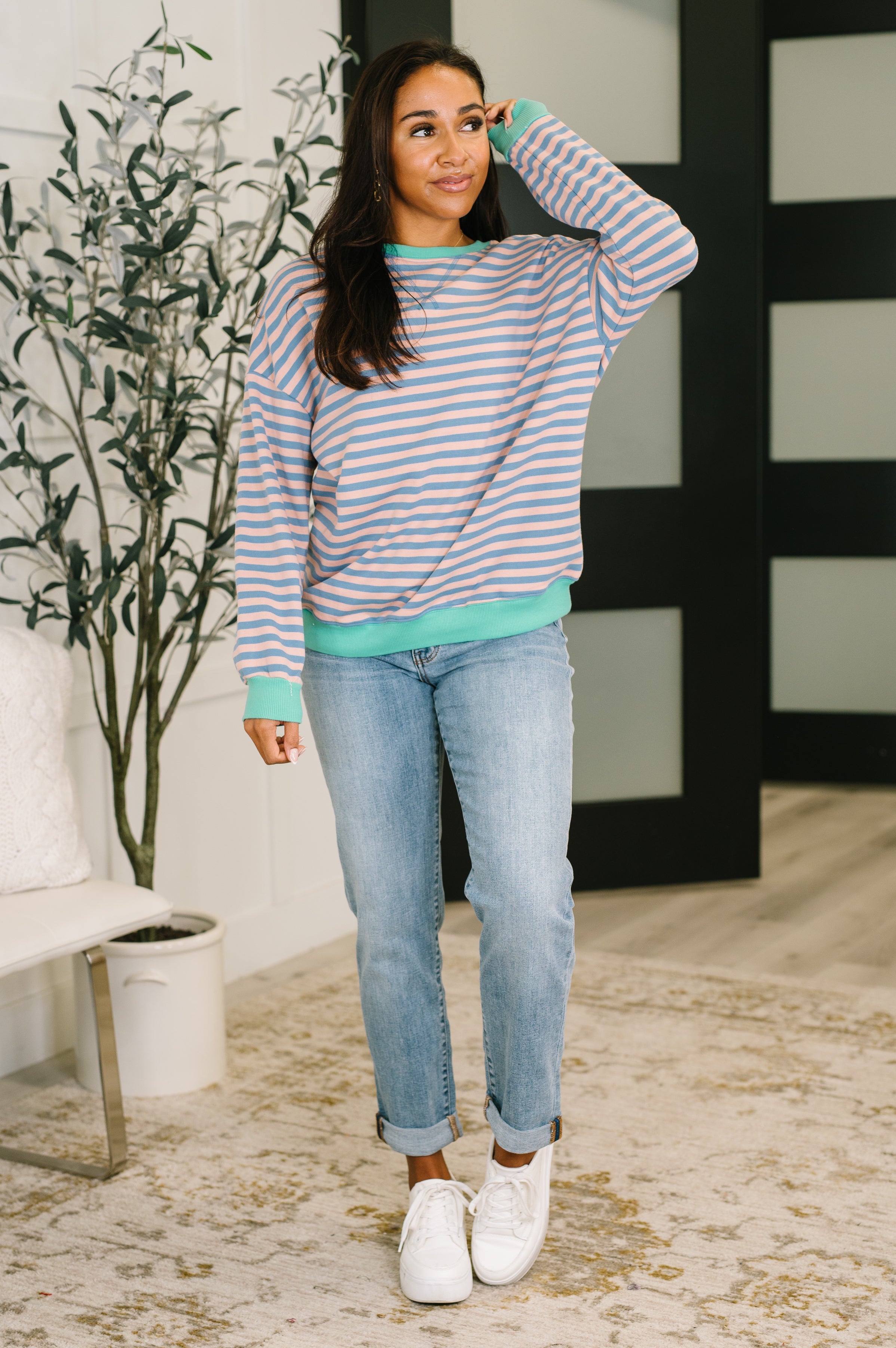 Woman wearing a peach and blue striped drop shoulder pullover, blue jeans and white sneakers standing in an indoor setting.