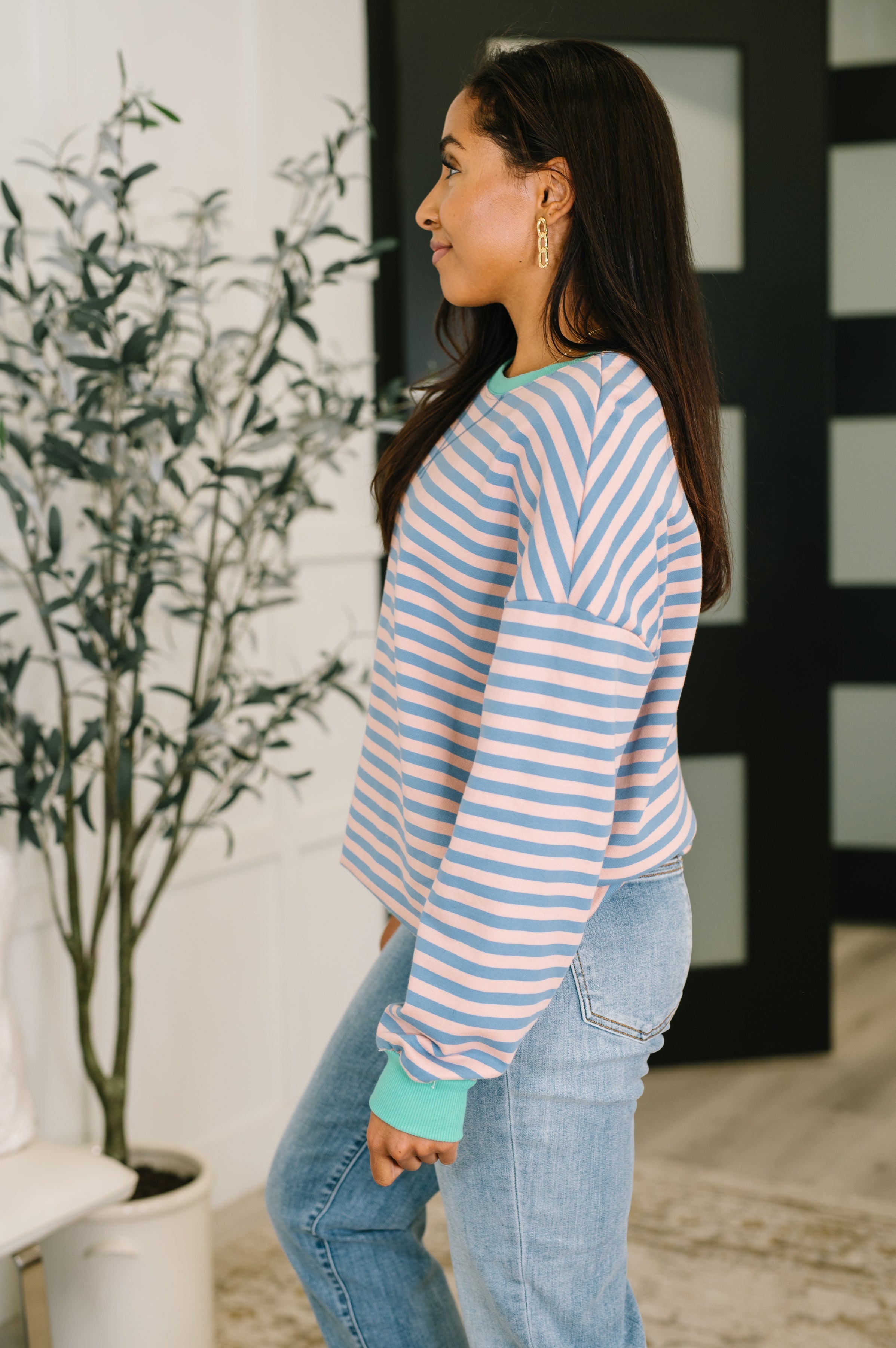 Woman wearing a peach and blue striped drop shoulder pullover and blue jeans standing in an indoor setting.