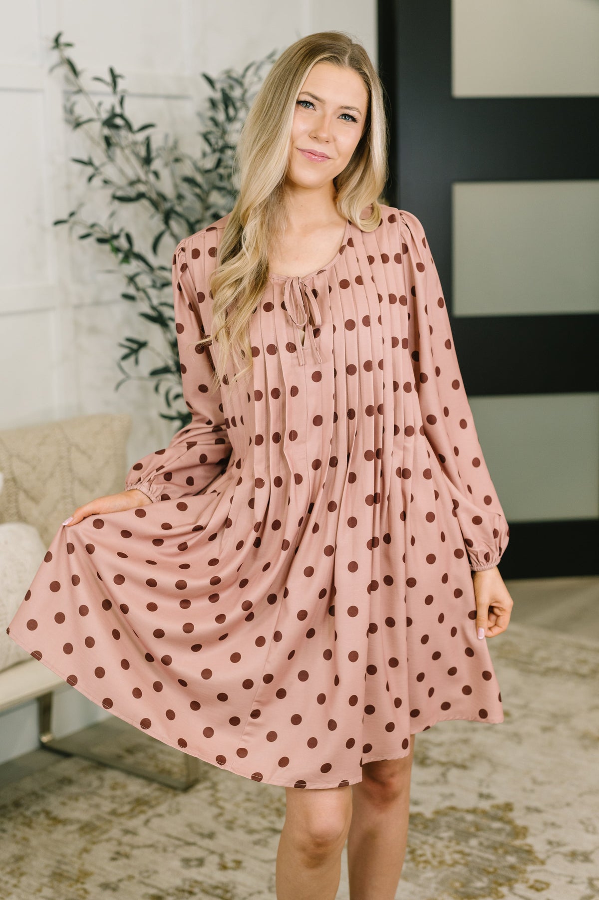 Woman models a pink pleated dress with brown polka dots and long sleeves while standing in a bright modern room