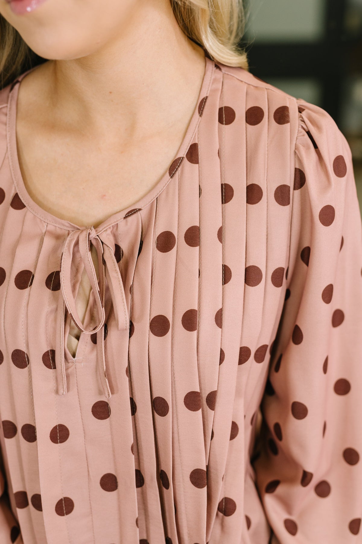 Close view of a woman wearing a pink pleated dress with brown polka dots puff sleeves and a tied scoop neckline