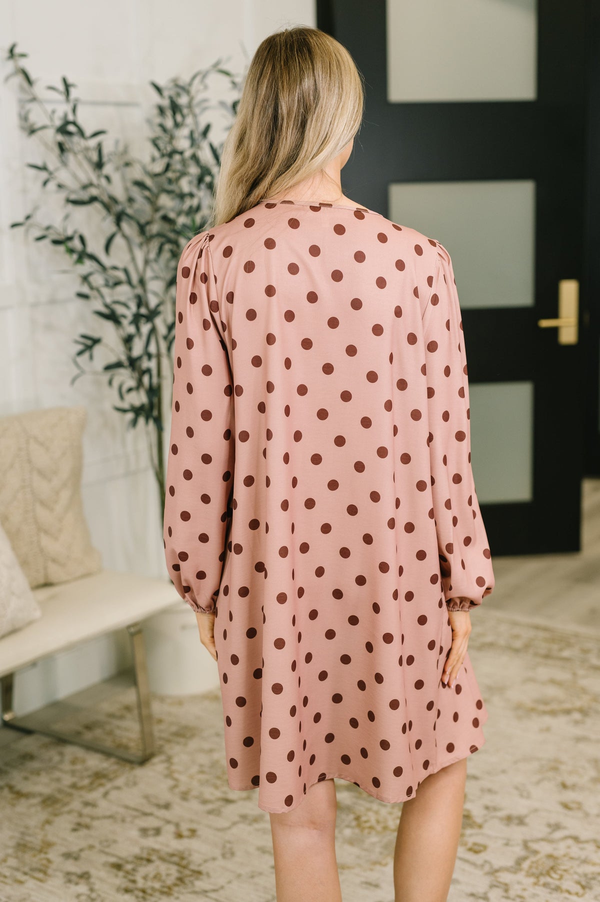 Back view of a woman modeling a pink pleated dress with brown polka dots and long sleeves in a bright modern room