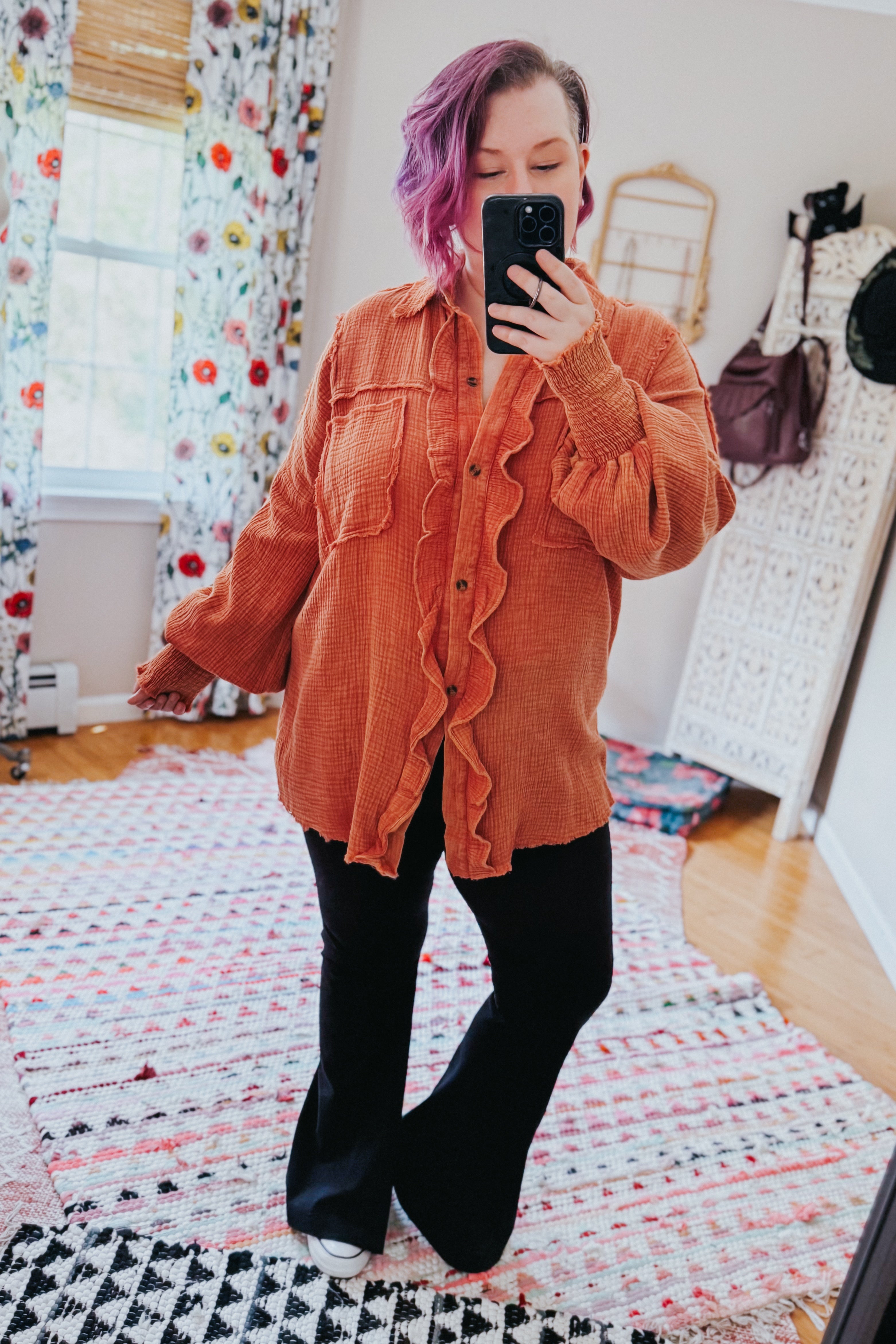 Woman with purple hair taking mirror selfie wearing loose orange ruffled button-up shirt with textured fabric and two chest pockets indoors