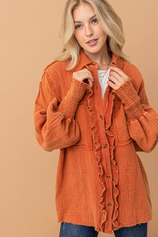 Young woman adjusting collar of textured burnt orange ruffled shirt with smocked cuffs against tan backdrop
