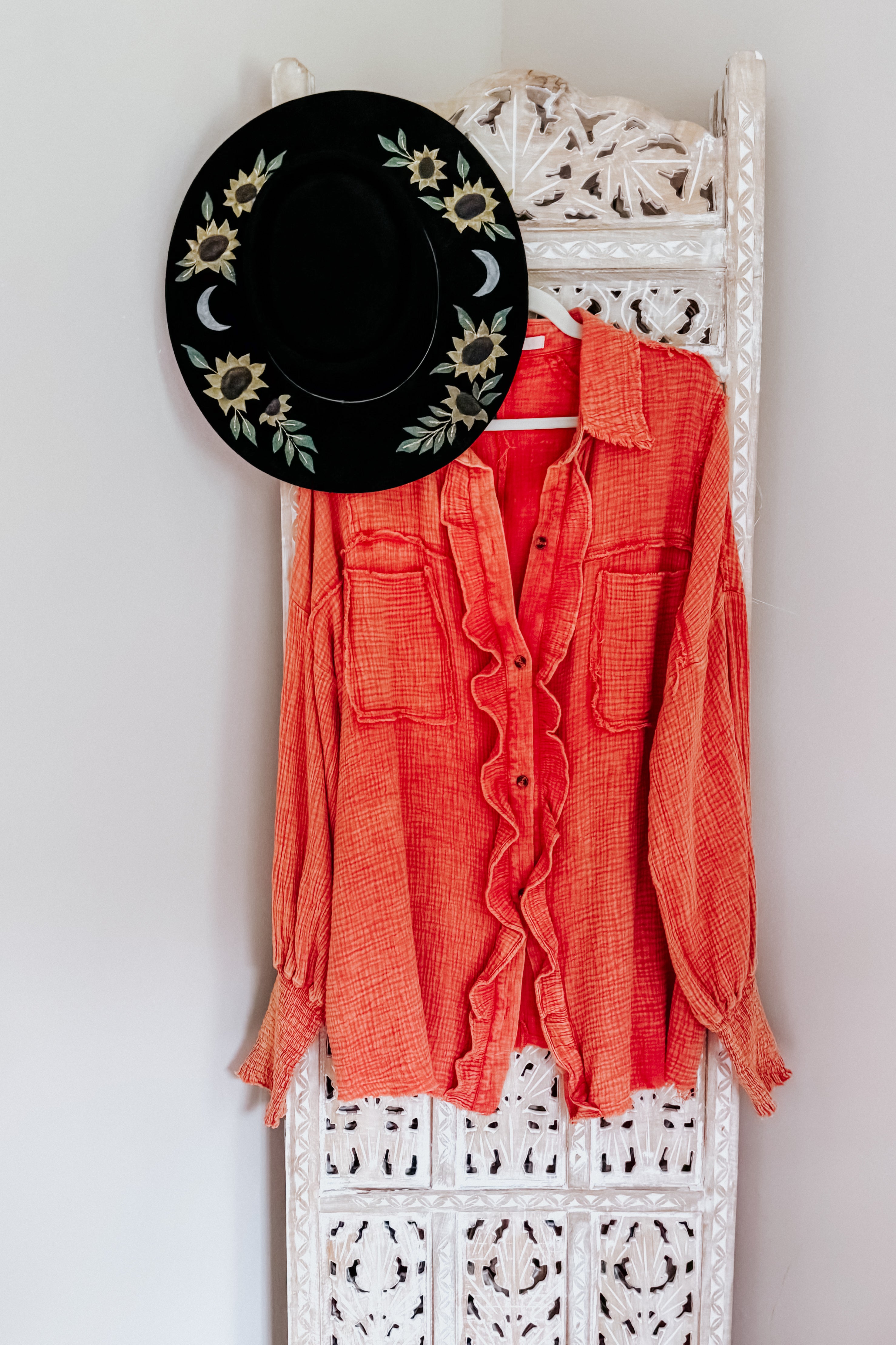 Orange textured All Pan Out Ruffled Button Up Shirt with front pockets hanging on carved white wooden panel with black floral hat
