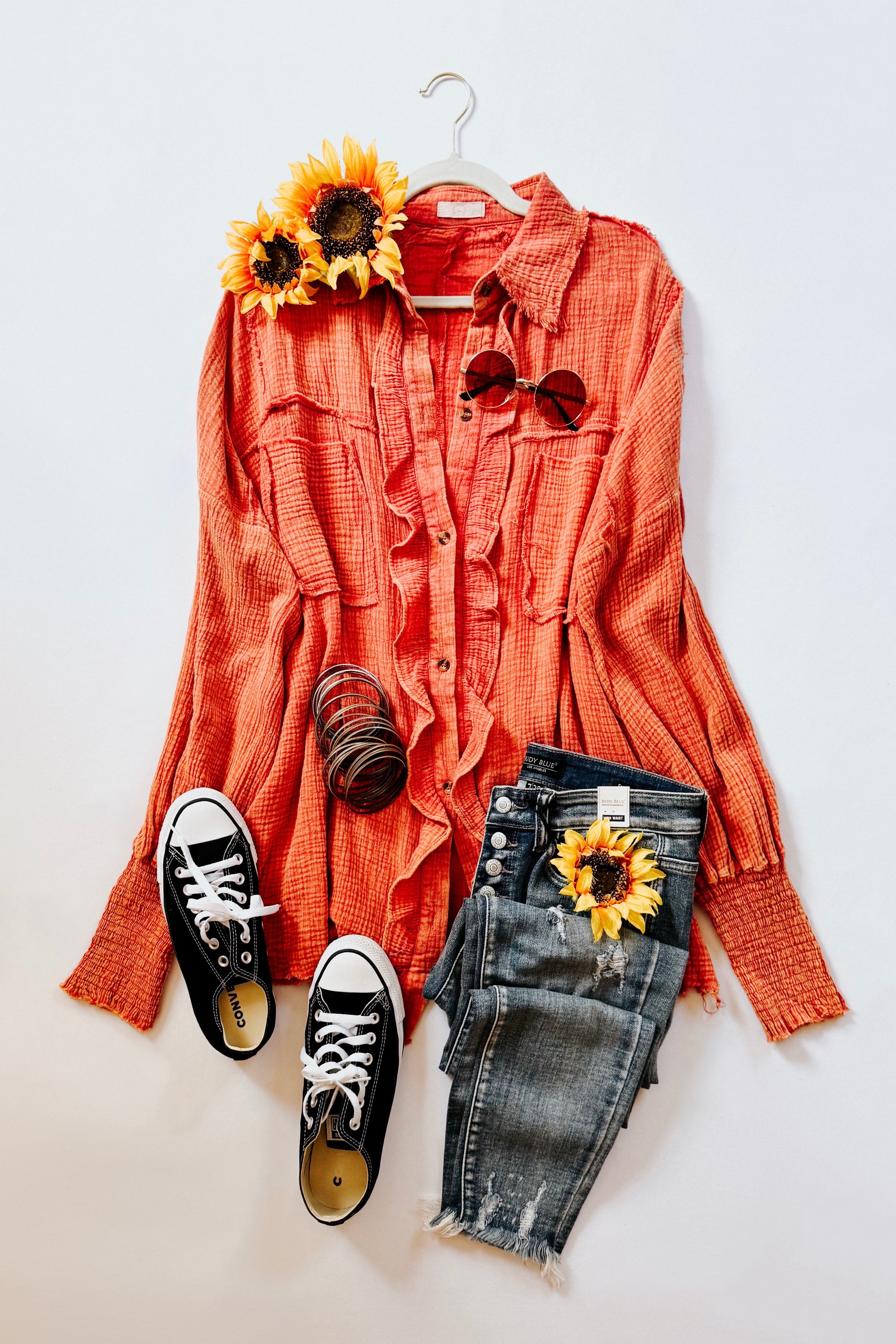 Orange ruffled button-up shirt styled flat with distressed jeans, black sneakers, sunglasses, bangles, and sunflowers on white background