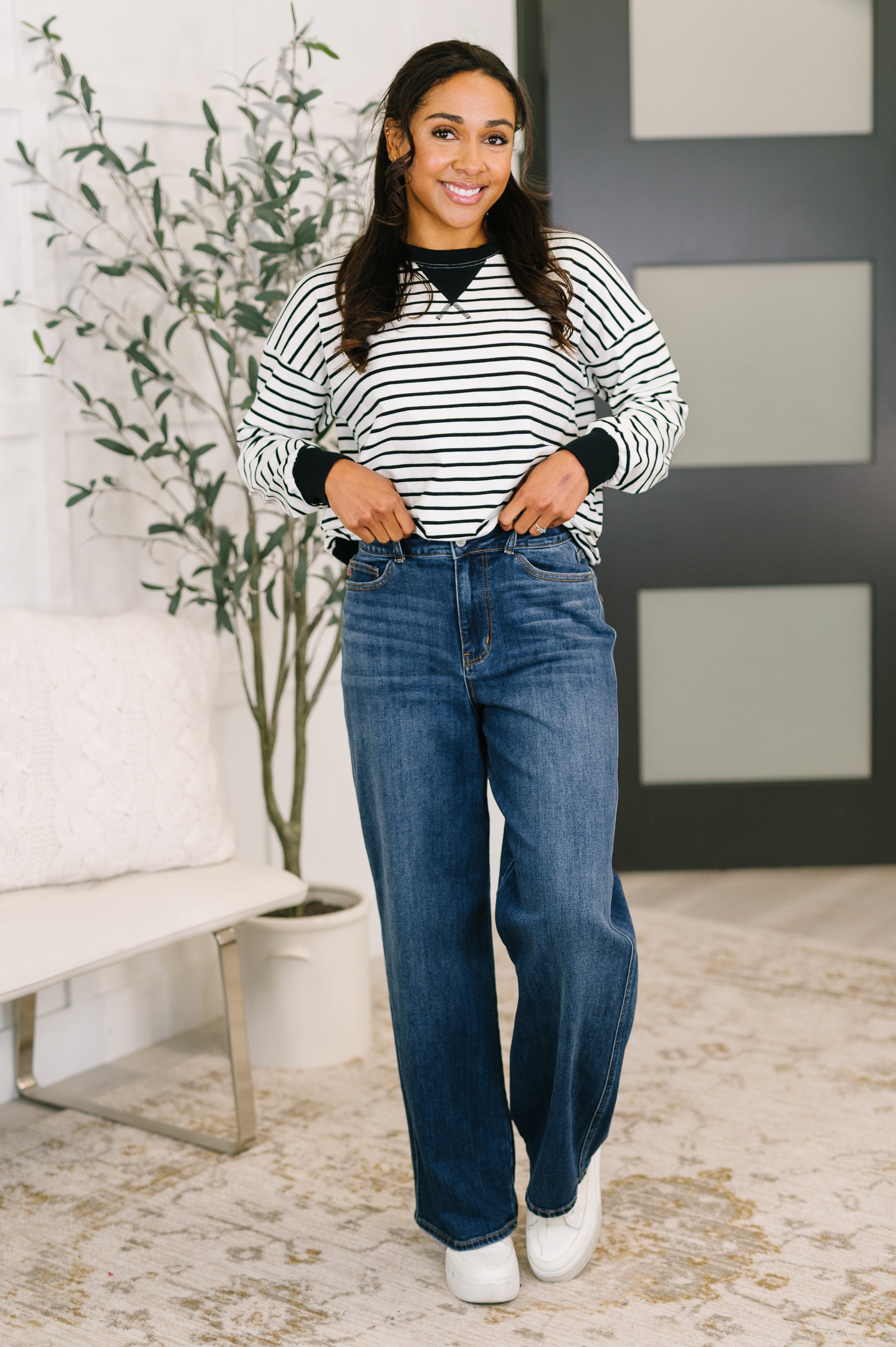 Smiling woman posing indoors wearing black and white striped Almost Therapeutic Drop Shoulder Pullover with blue jeans and white shoes