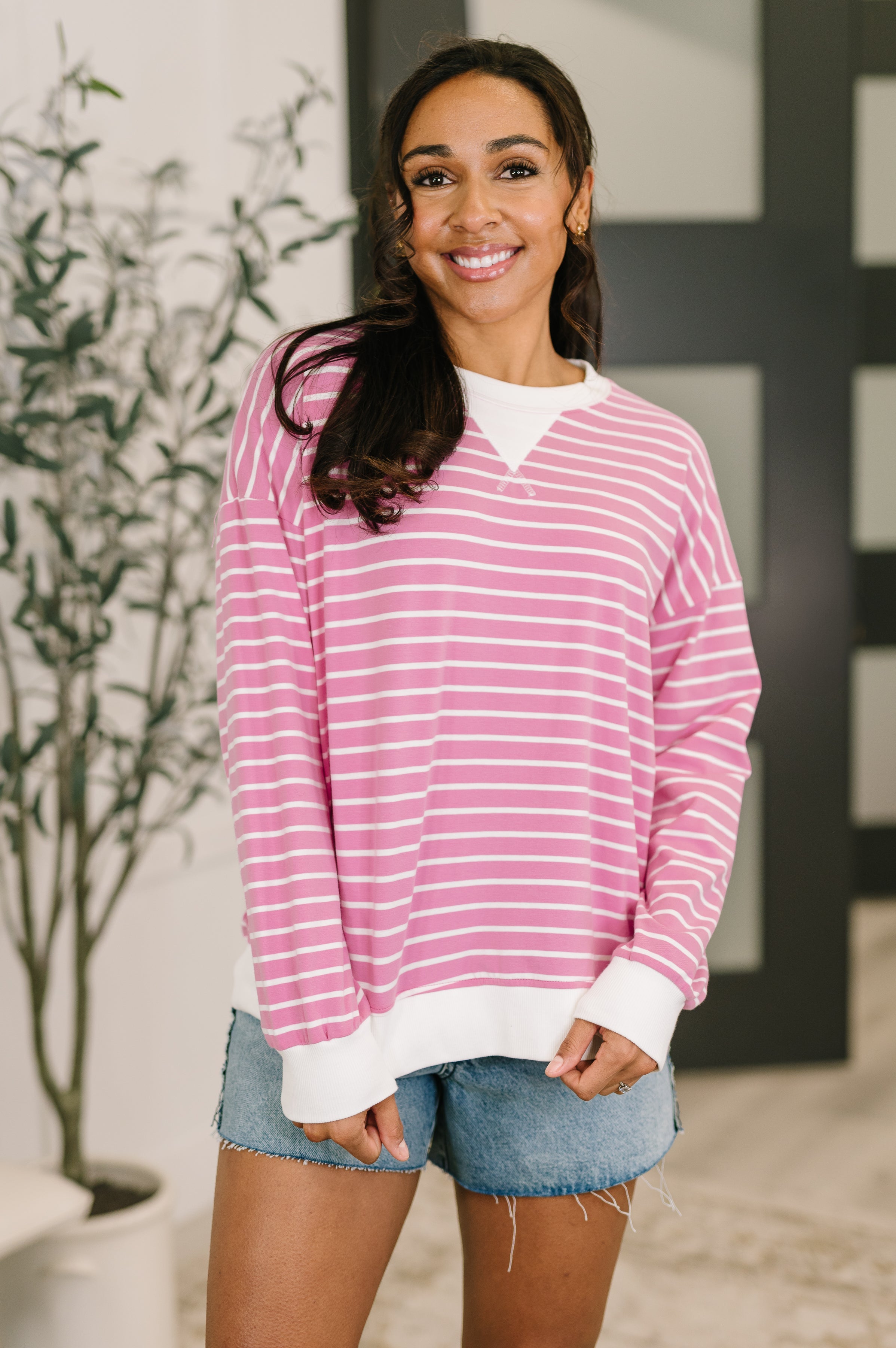 Smiling woman indoors wearing candy pink Almost Therapeutic Drop Shoulder Pullover with white stripes and cuffs paired with denim shorts