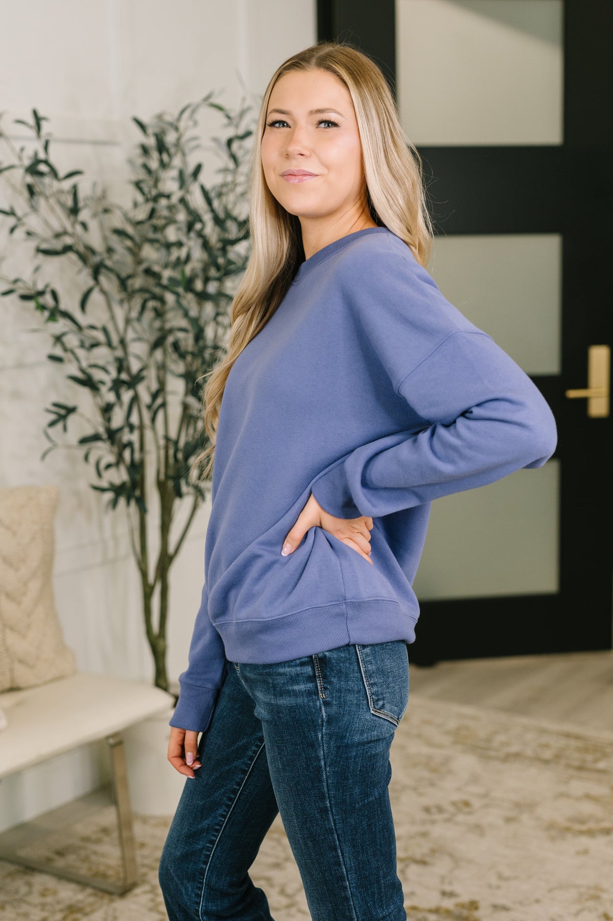 Woman models a blue drop shoulder pullover with a kangaroo pocket and jeans in a softly styled modern home interior