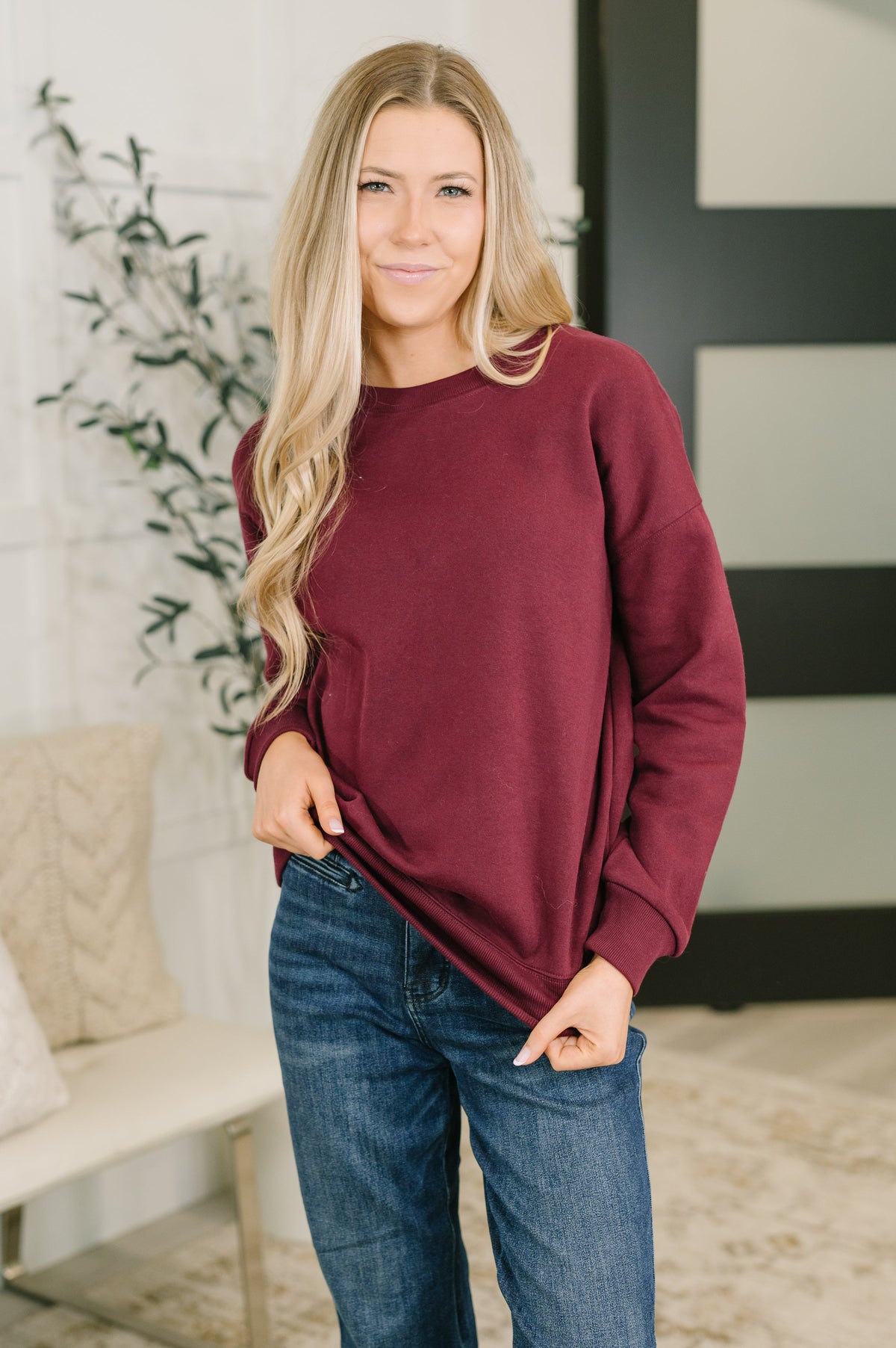 Woman modeling a burgundy drop shoulder pullover with relaxed sleeves and blue jeans in a bright modern home
