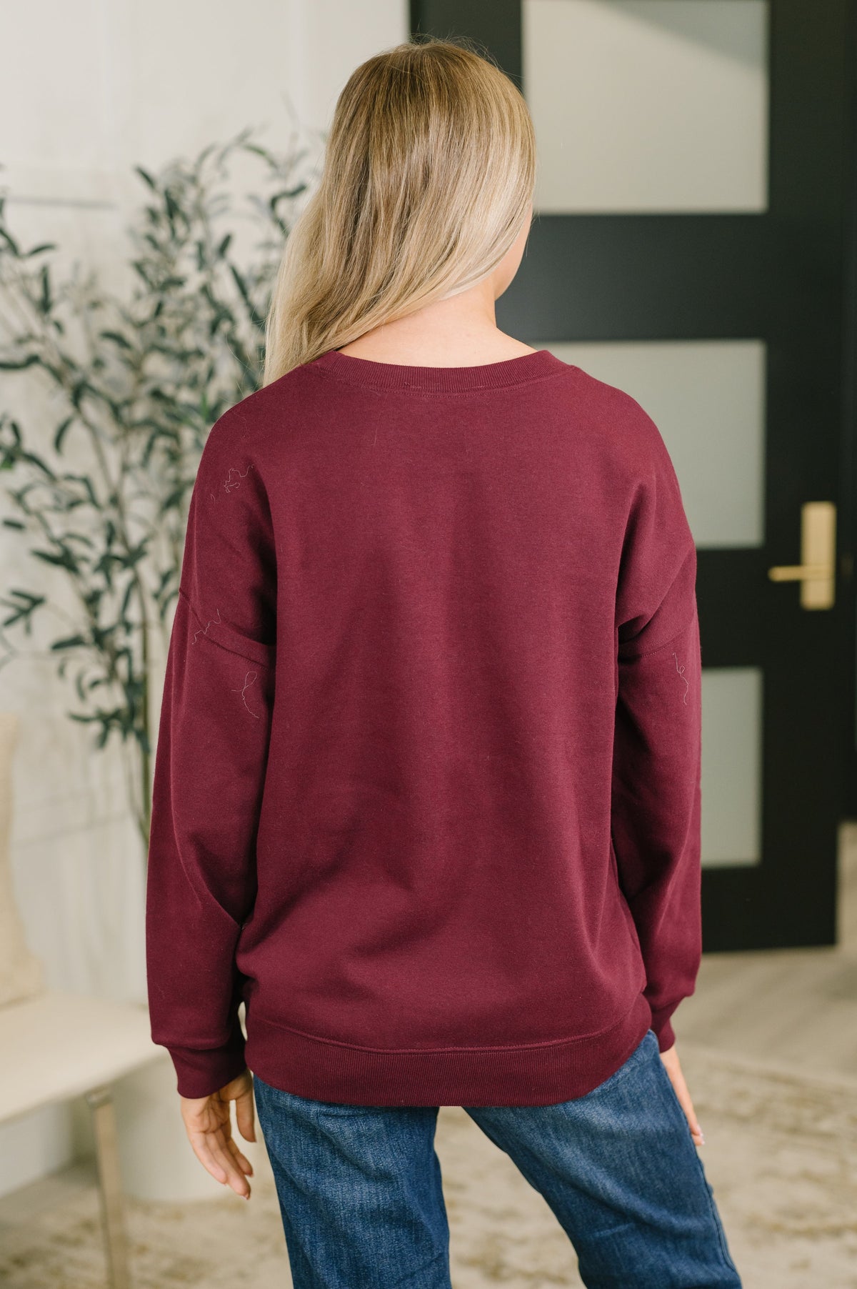 Woman seen from behind wearing a dark burgundy drop shoulder pullover with blue jeans in a bright home entryway