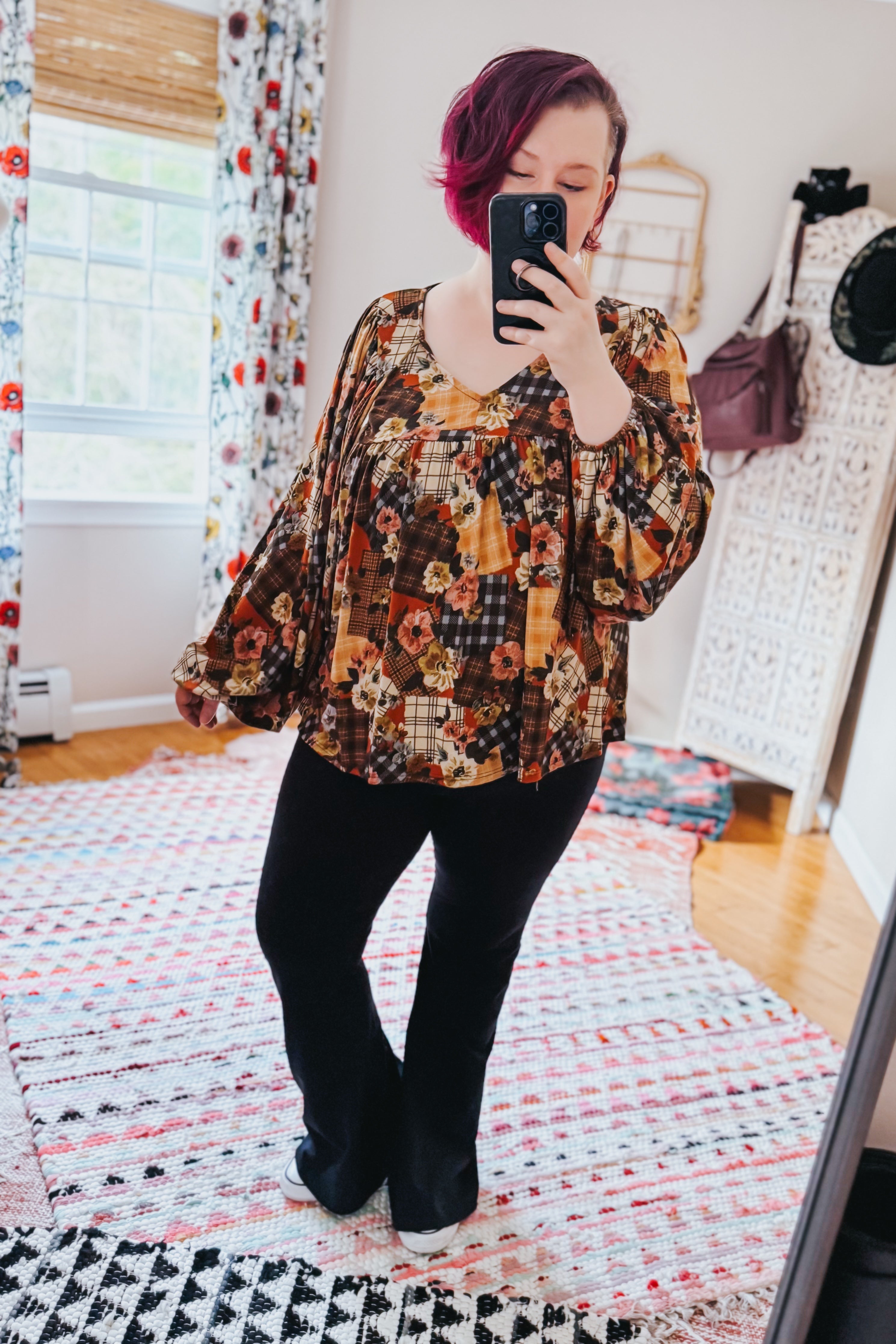 Woman taking a mirror selfie wearing a patchwork print blouse and black pants in a room with colorful curtains and a rug.