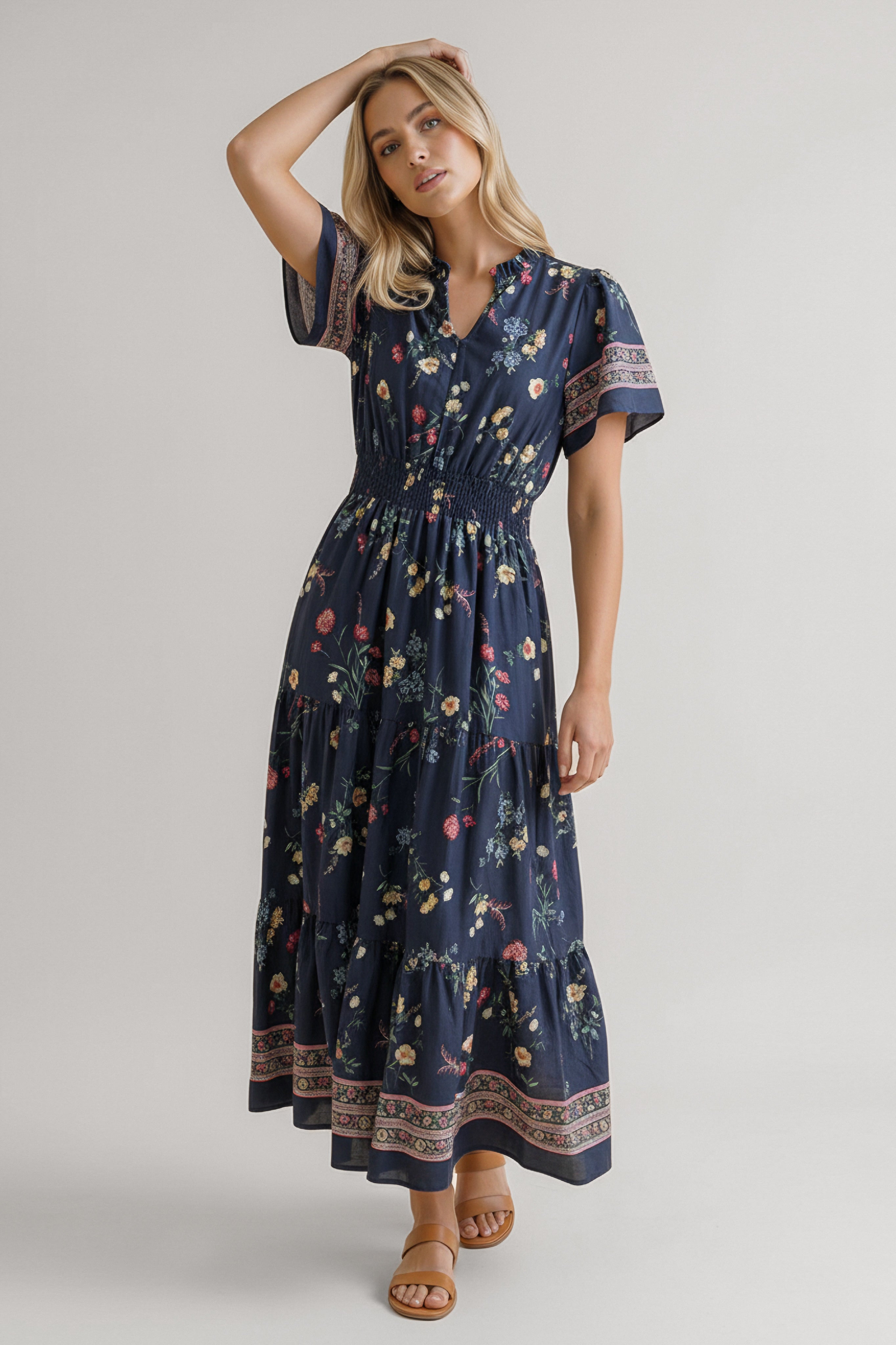 Blonde woman wearing a navy floral dress with short sleeves, smocked waist, and patterned borders standing barefoot