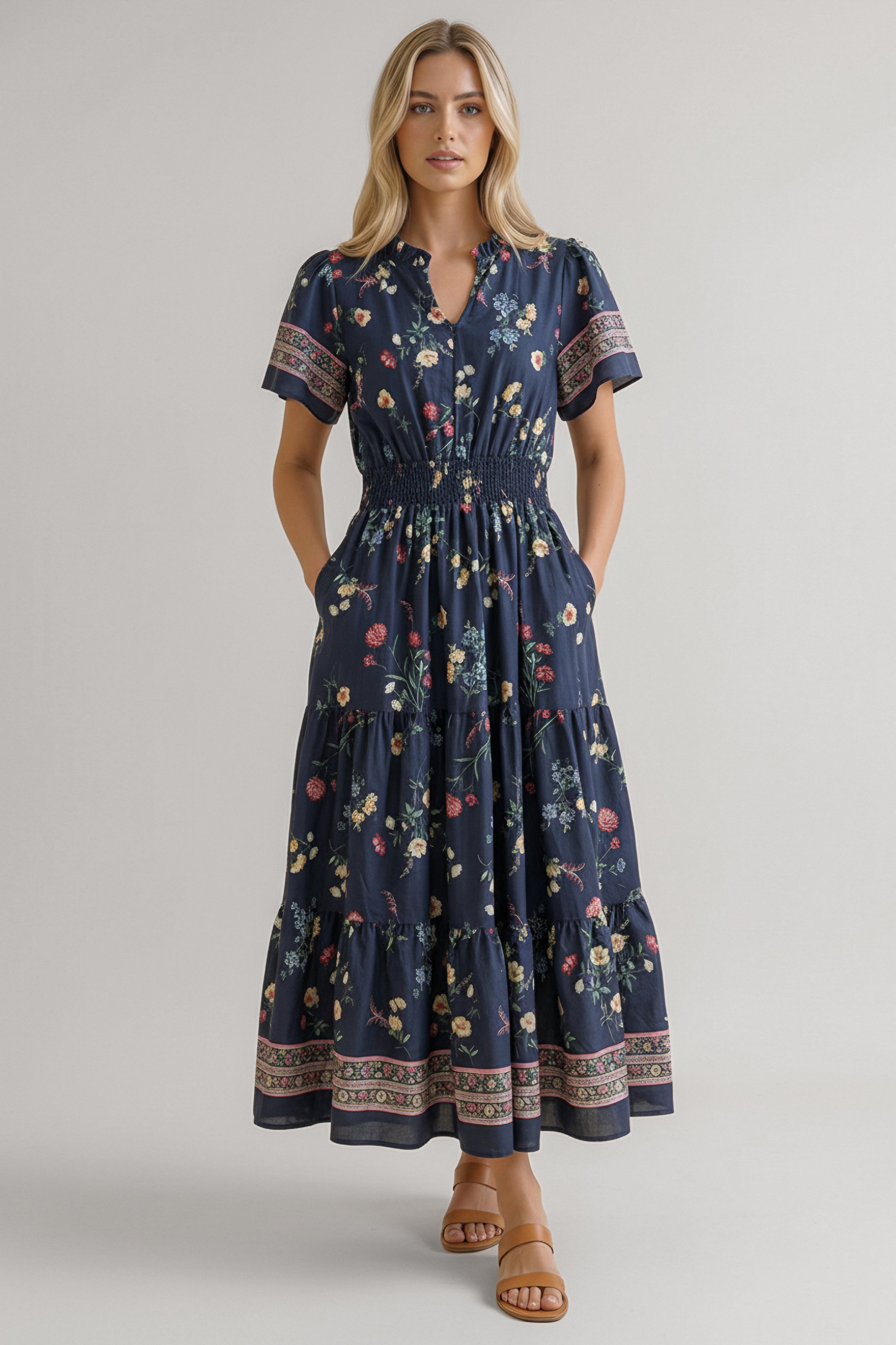 Blonde woman wearing a navy floral midi dress with smocked waist, short sleeves, and patterned borders standing with hands in pockets