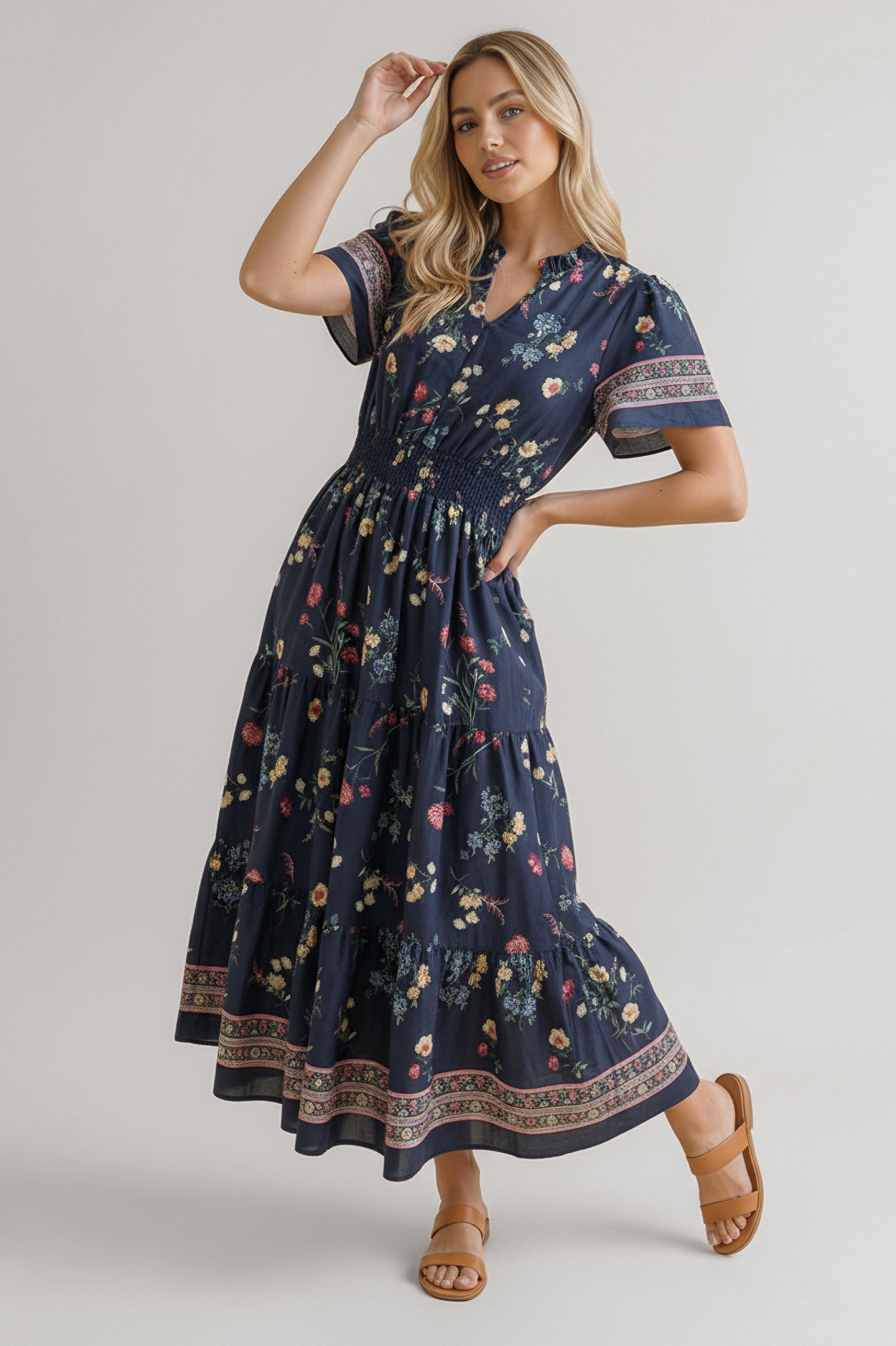 Woman wearing a navy floral maxi dress with smocked waist and patterned borders posing with one arm raised indoors
