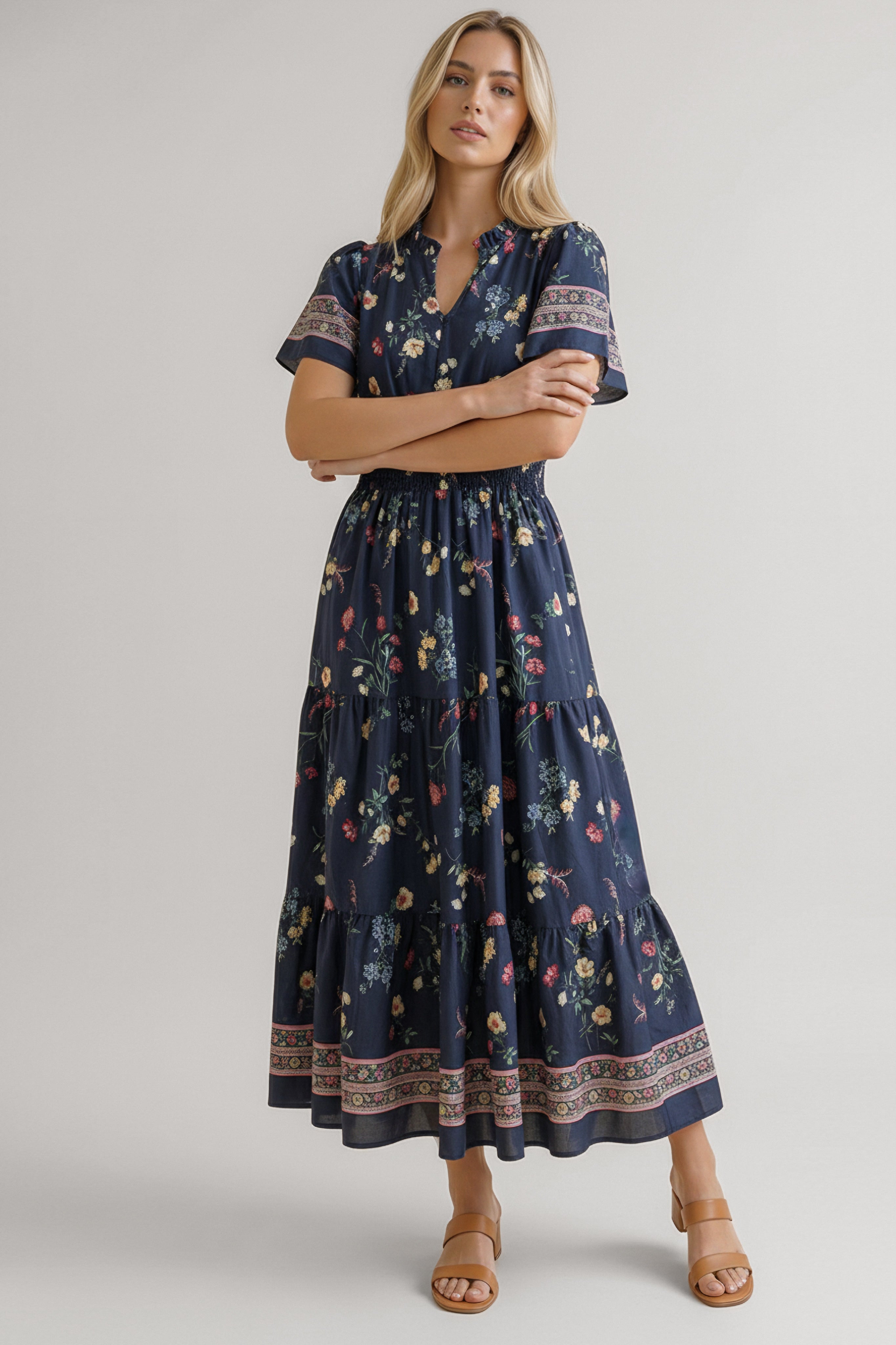 Blonde woman wearing a navy floral dress with short sleeves and patterned trim standing with arms crossed indoors