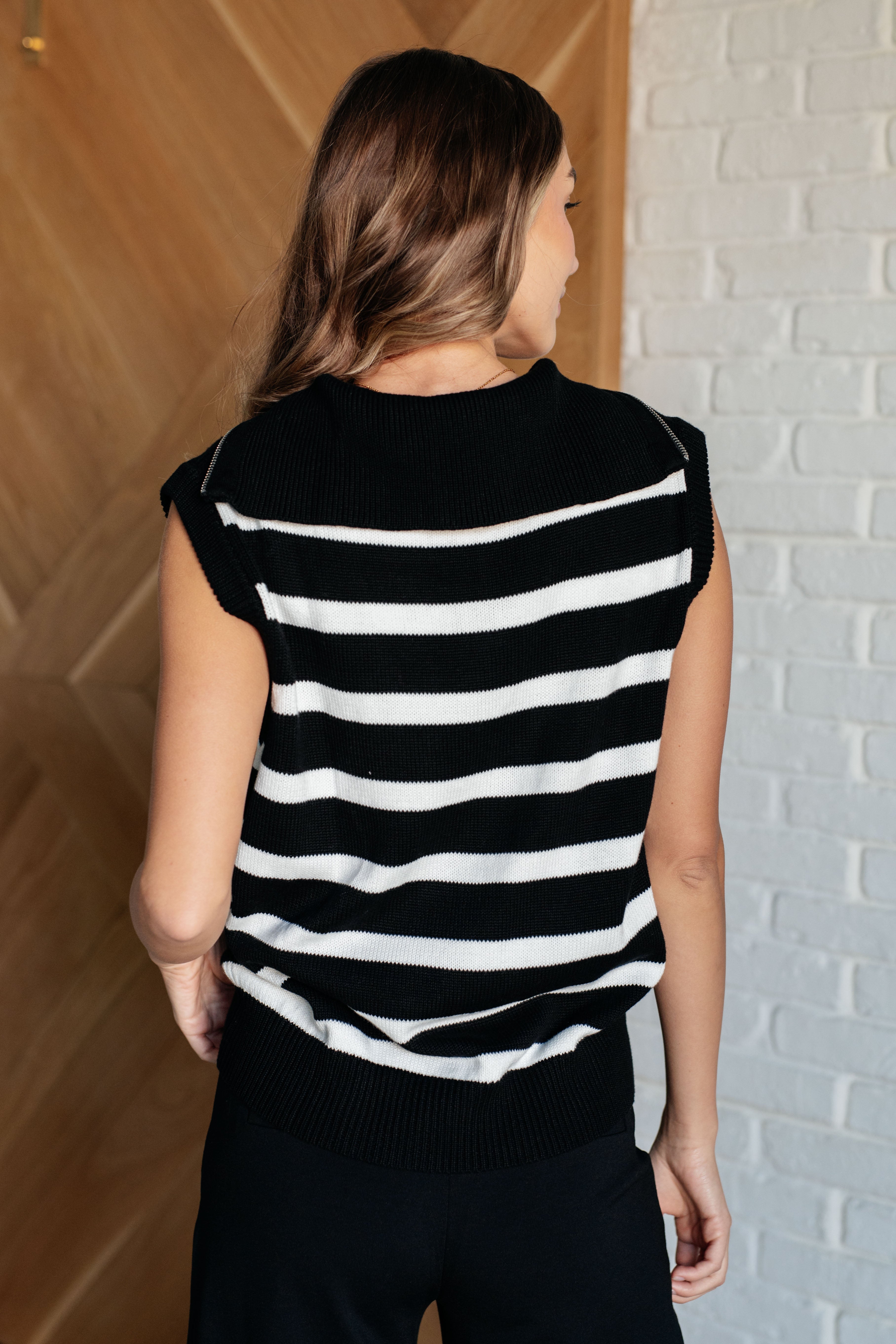 Rear view of woman wearing black and white striped sleeveless 1/4 zip sweater with ribbed hem and neckline