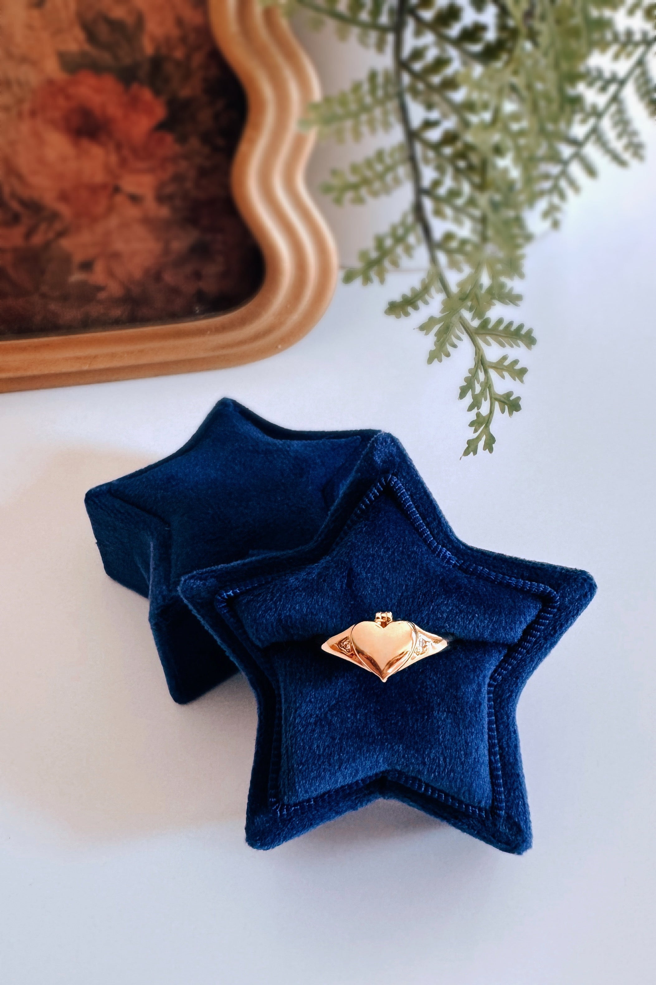 Gold heart-shaped locket ring displayed inside a blue velvet star-shaped ring box on white surface with greenery nearby