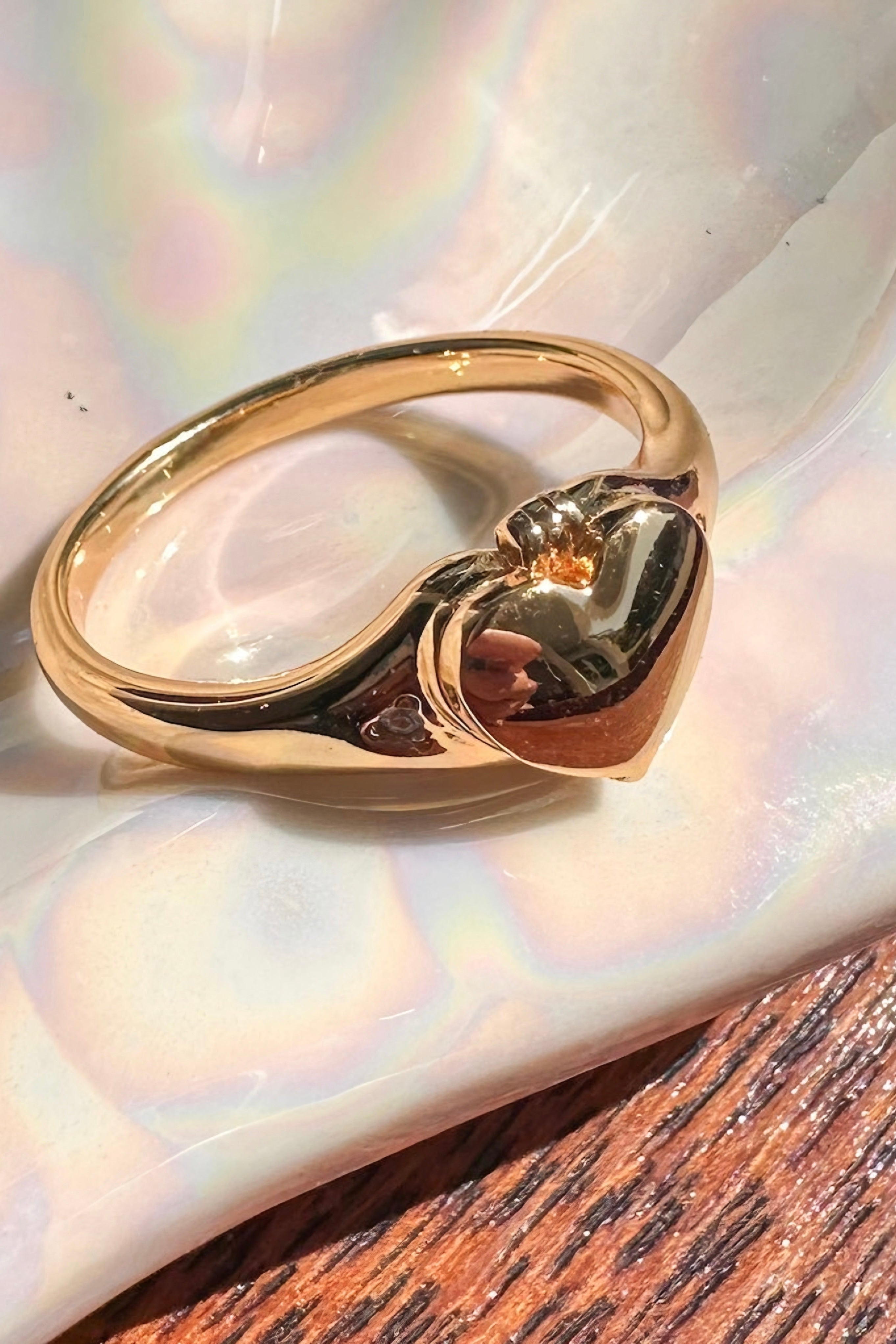 Gold heart locket ring with smooth, polished surface resting on iridescent shell and wooden background