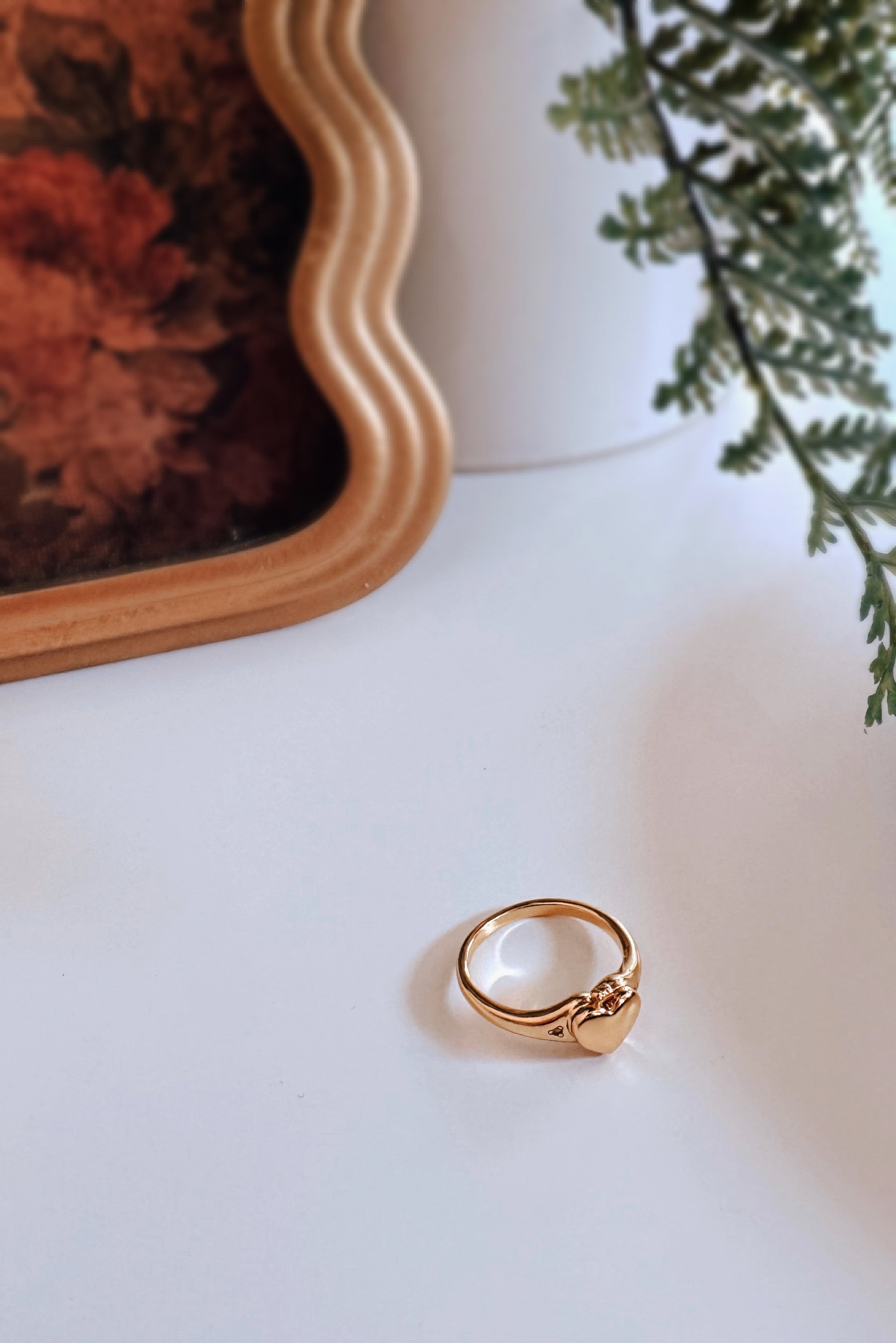 Gold Aurelia Heart Locket Ring on white surface with blurred wavy mirror and green foliage in background