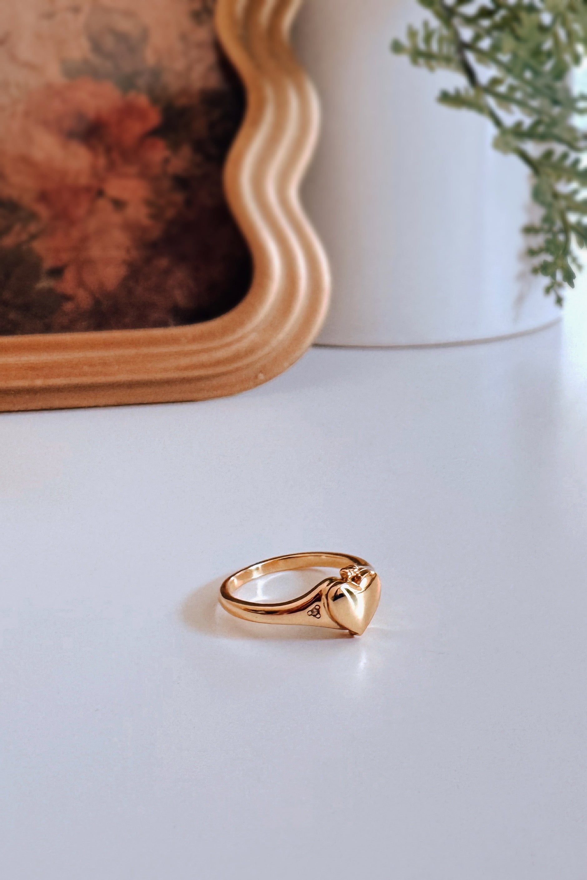 Gold Aurelia Heart Locket Ring with smooth heart-shaped locket on white surface by framed floral artwork and greenery