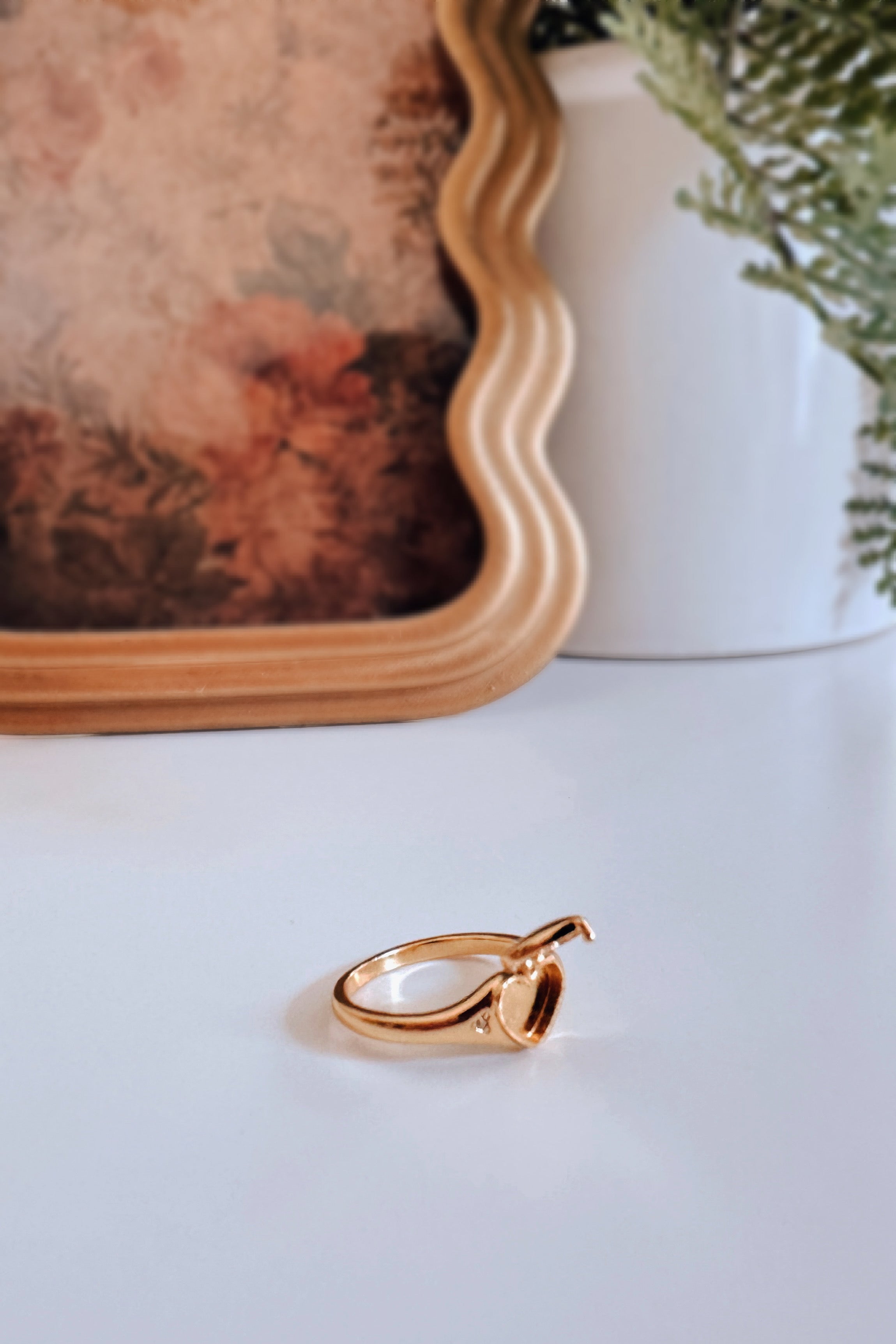 Gold Aurelia Heart Locket Ring open to show heart-shaped compartment on white surface with blurred floral art and greenery background