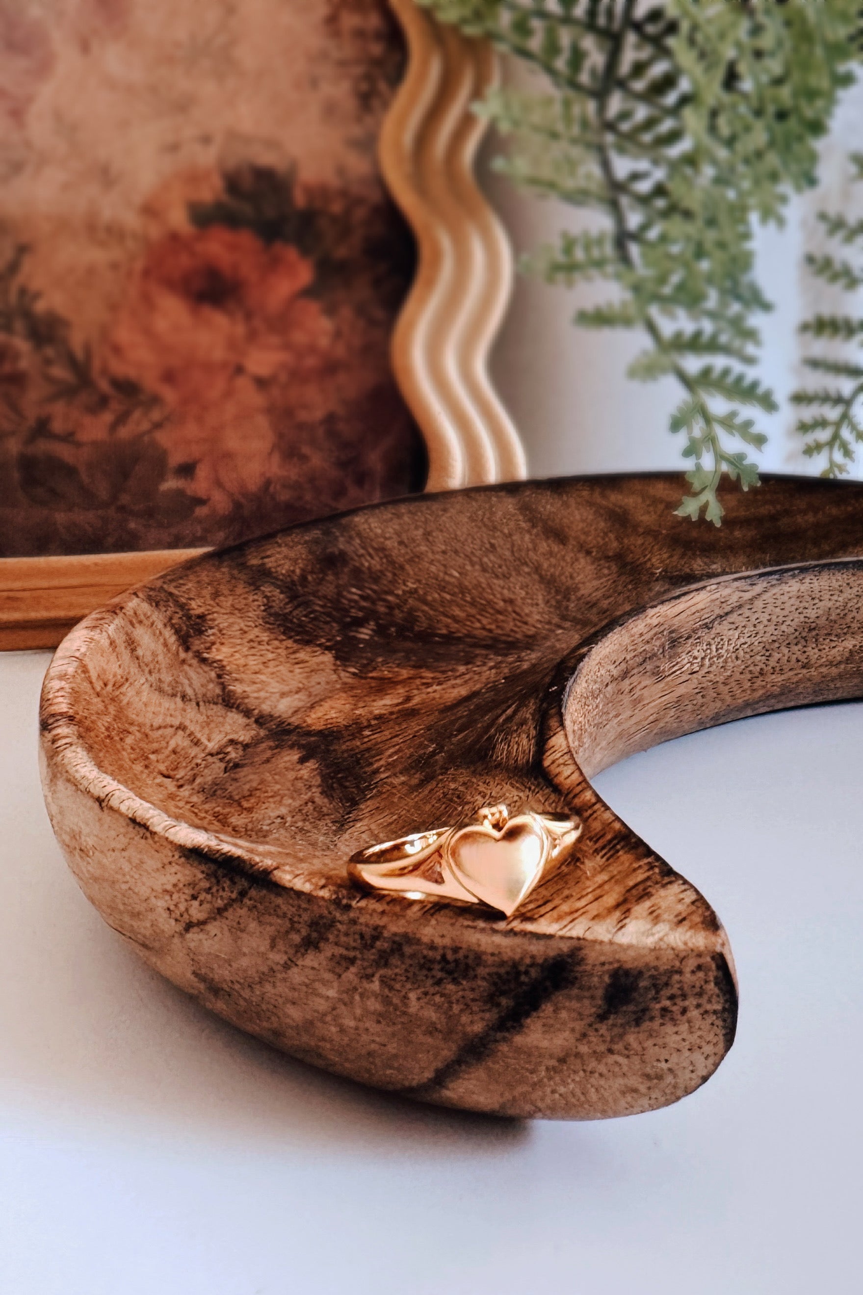 Golden Aurelia Heart Locket Ring displayed on a curved wooden tray with greenery and framed artwork nearby