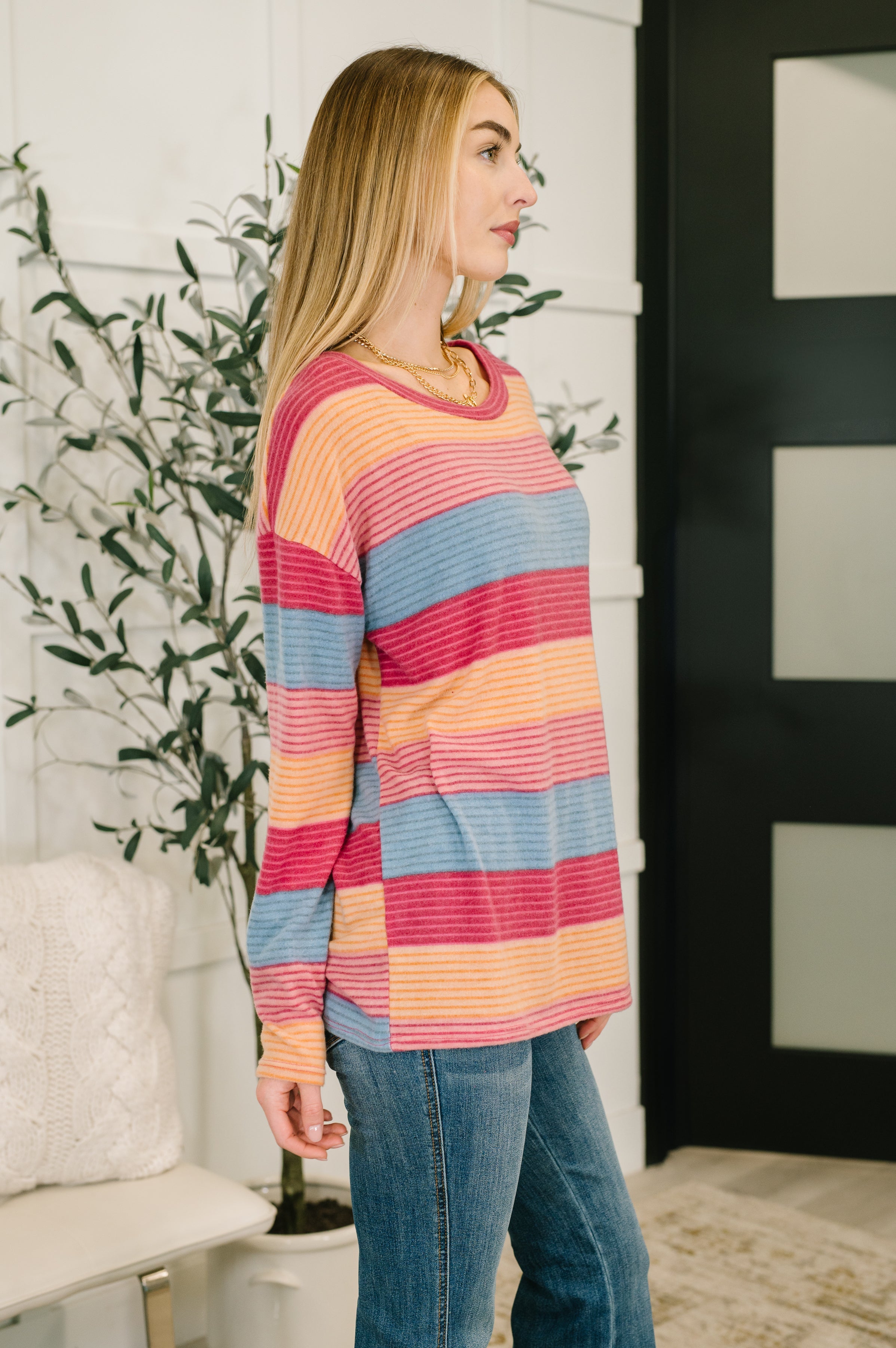 Woman wearing a blue, orange and red striped drop shoulder long sleeve top, a gold necklace and blue jeans in an indoor setting.