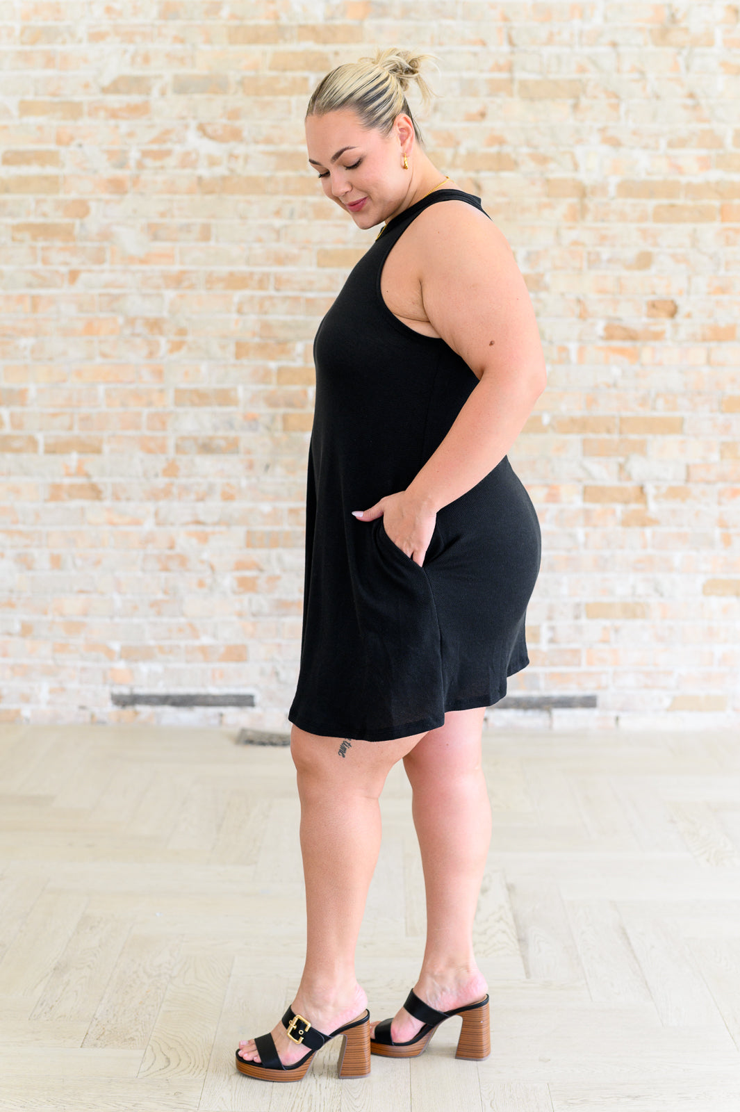 Plus-size woman in black sleeveless dress with pockets and wooden block heel sandals against brick wall