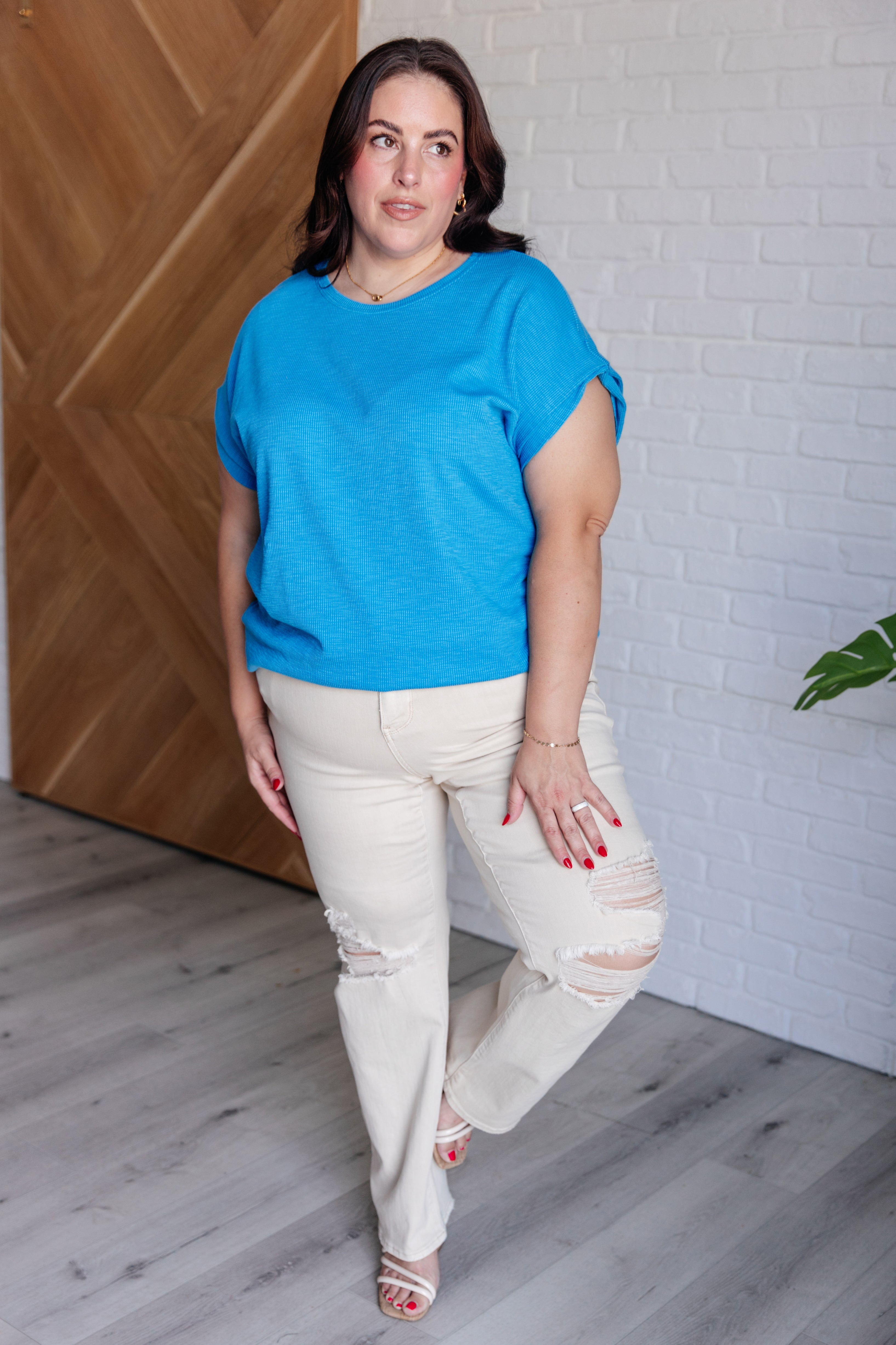 Plus size woman wearing bright blue short sleeve top and distressed cream jeans, standing indoors near wood door angle